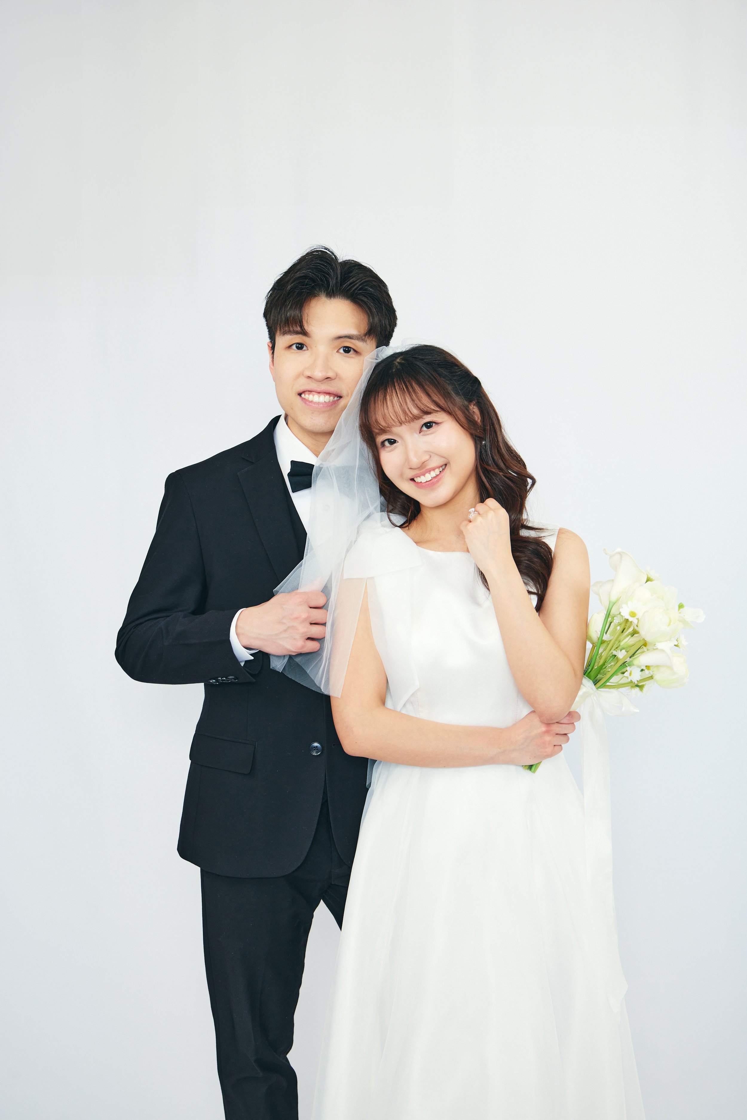 A happy bride and groom standing together, with the groom in a black tuxedo and the bride in a white wedding dress holding a bouquet, against a plain white background.