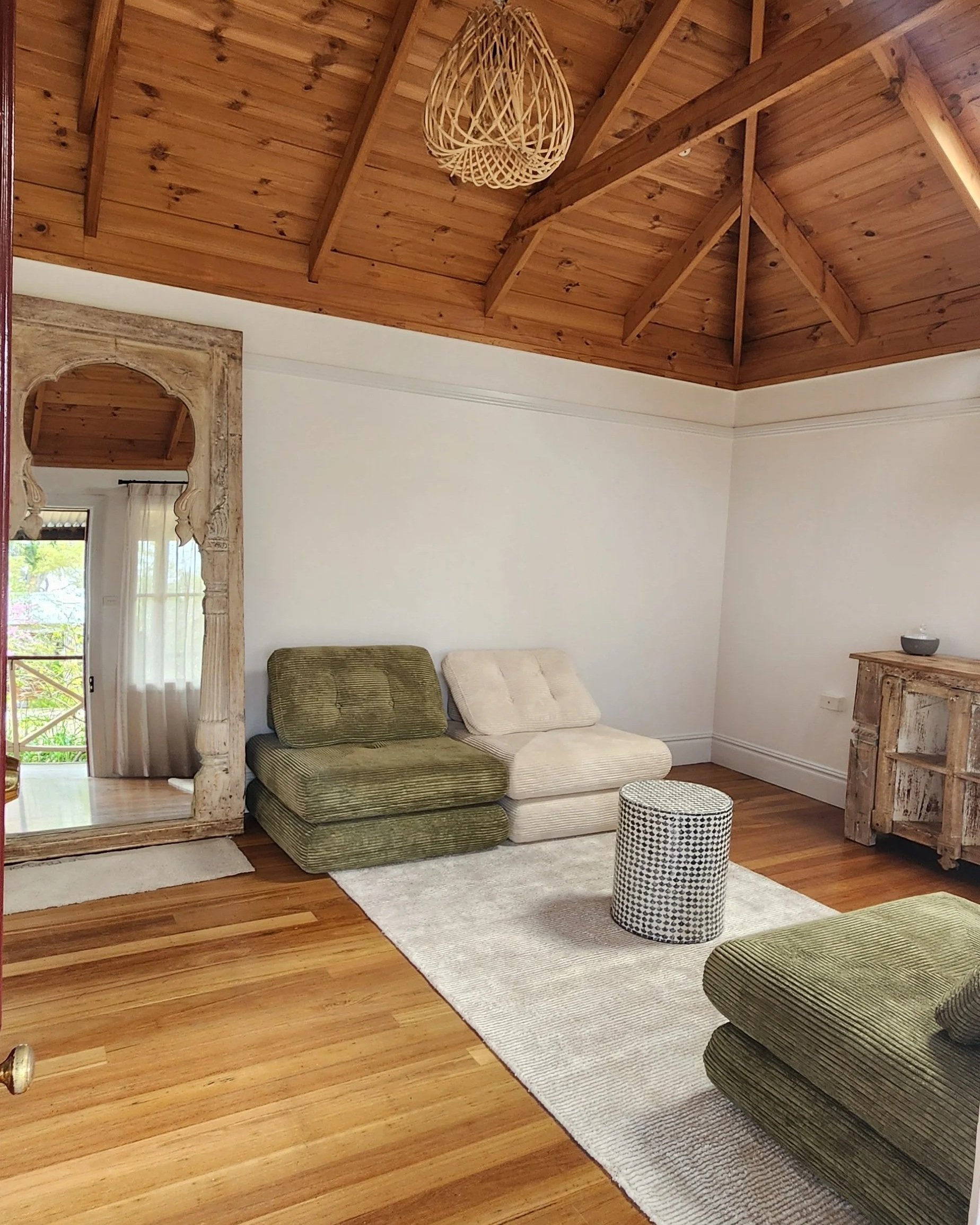 Living room with a wooden ceiling, white walls, a mirror, a green couch, a beige couch, a round patterned ottoman, a wooden cabinet, and a white rug.