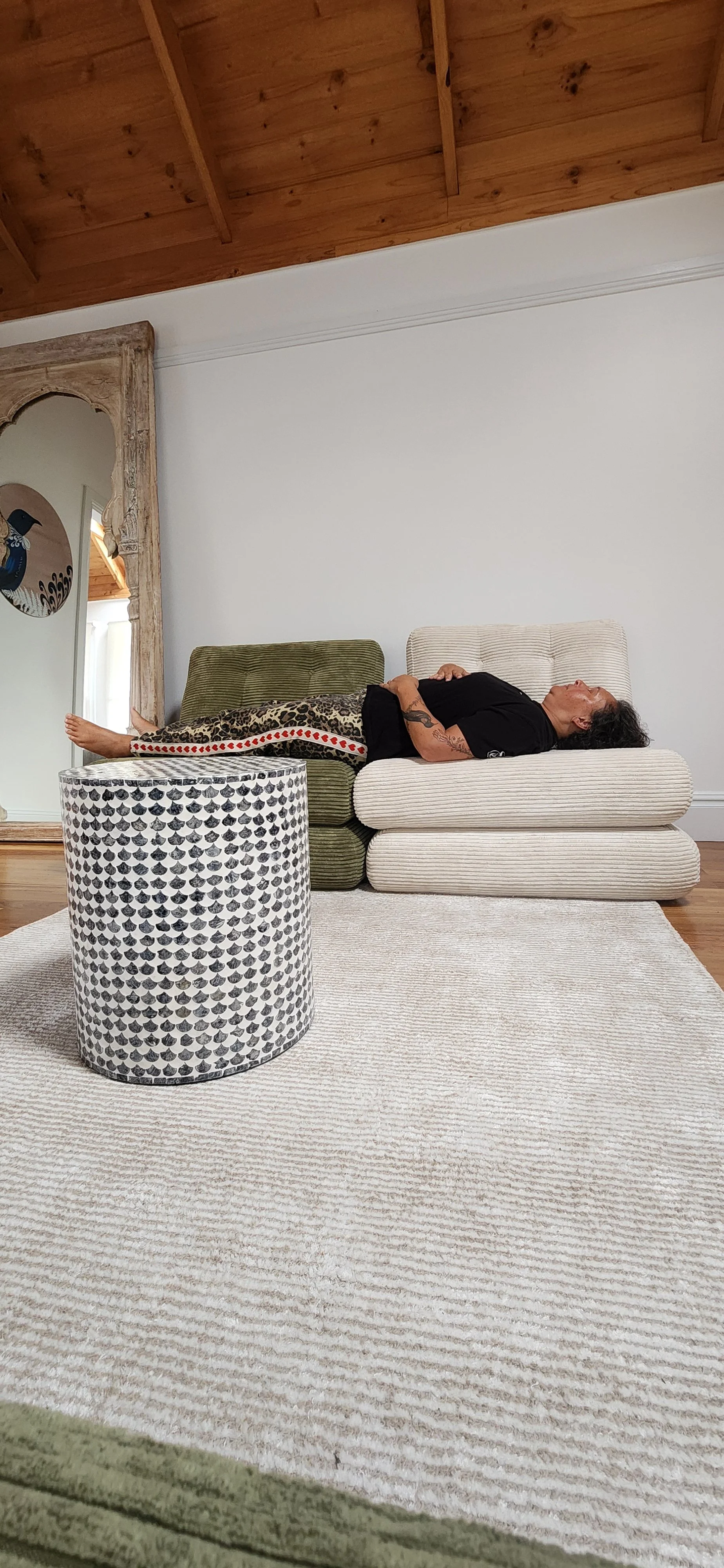A person lying on a large, layered beige cushion on the floor, with their head resting on the cushion and their legs stretched out across a green cushion. They are wearing a black shirt and patterned pants, with their eyes closed, appearing relaxed. There is a patterned cylindrical ottoman or side table in front of them, and a large mirror with a rustic wooden frame leaning against the wall behind. The room has wooden ceiling beams and a light-colored rug on the floor.
