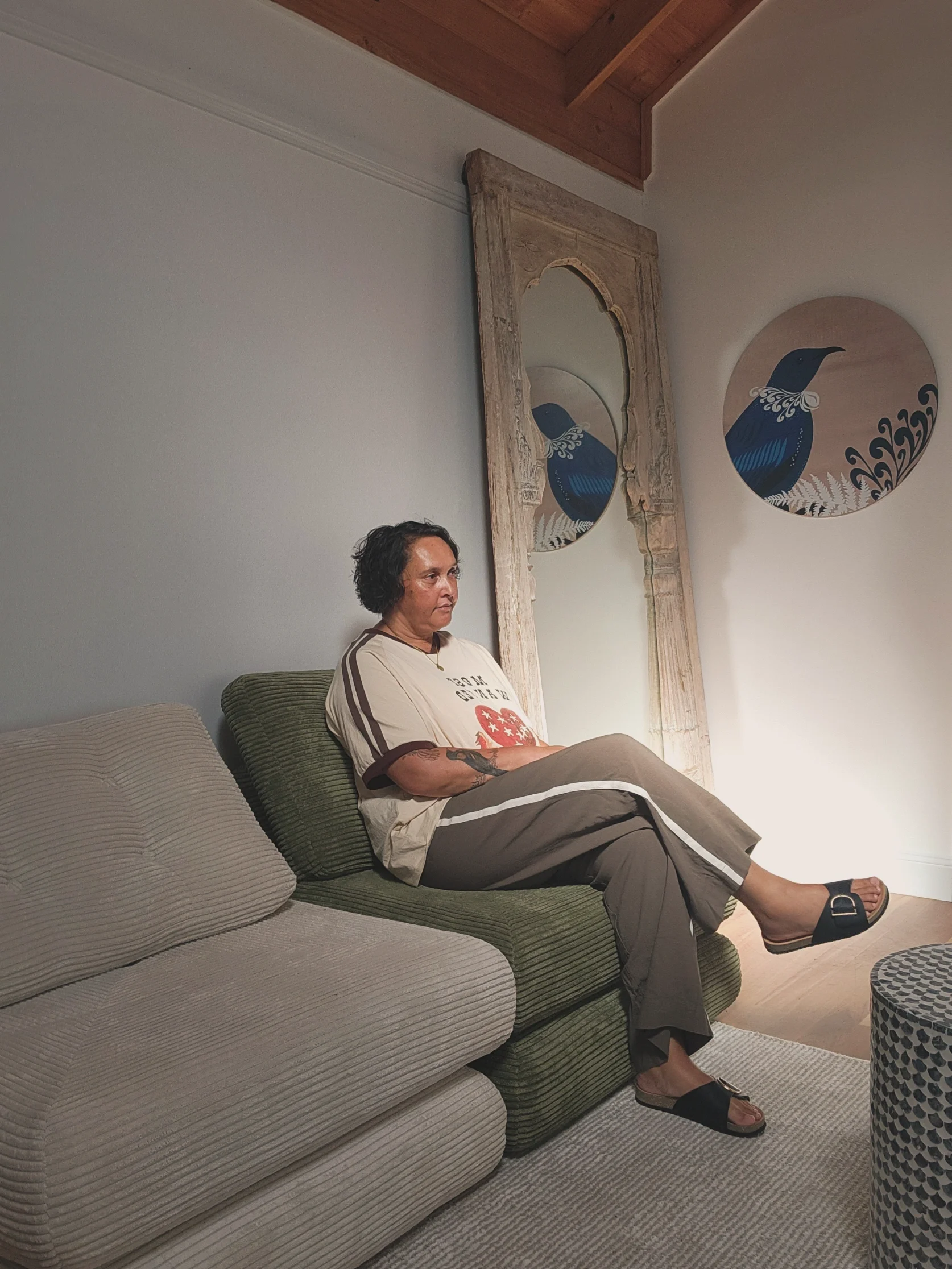 A woman sitting on a green sofa in a living room with art on the wall, including a round piece with a bird design and a tall mirror with a wooden frame.