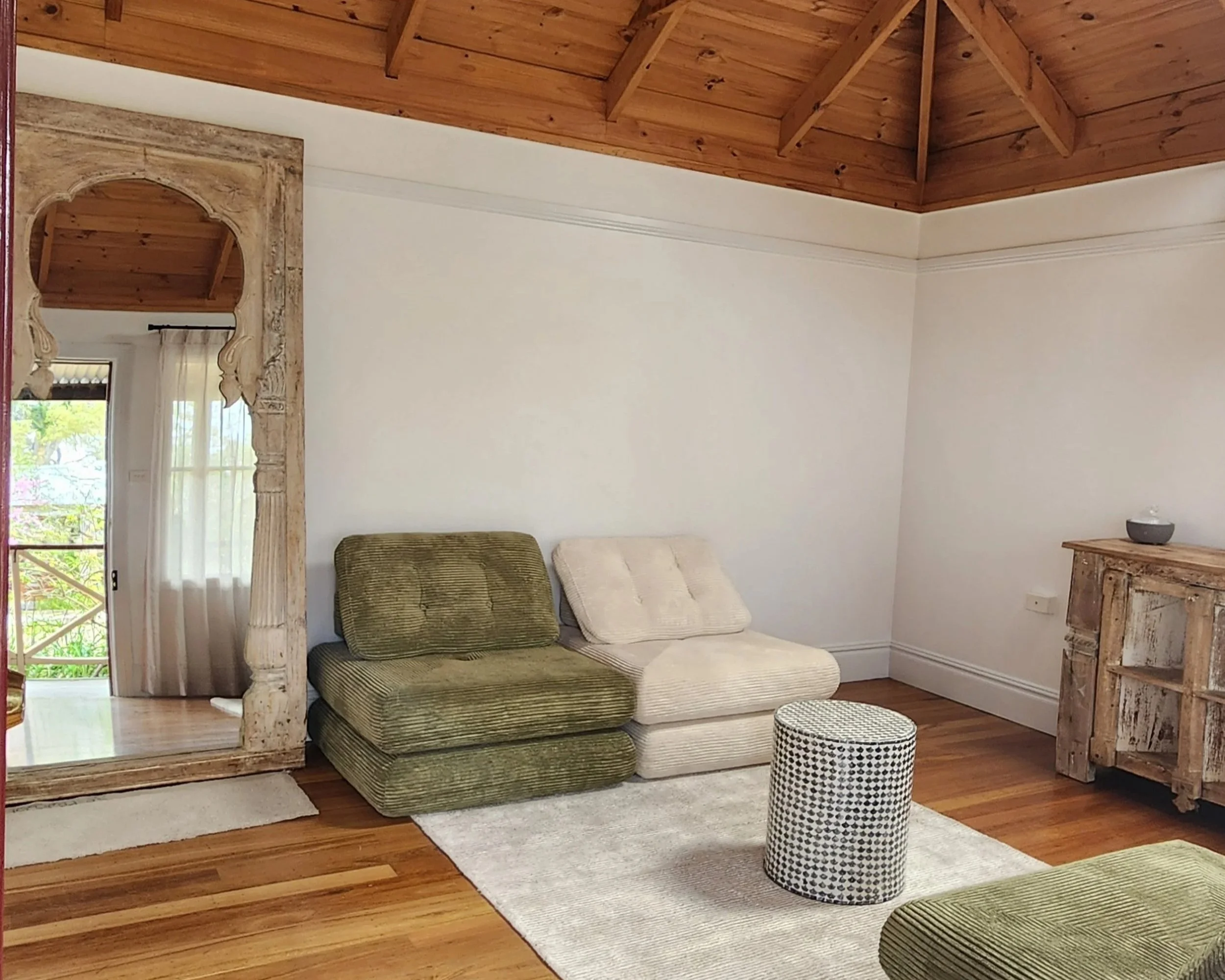 Living room with wooden ceiling, white walls, a large mirror with carved frame, a beige and green couch, a patterned cylindrical stool, a wooden cabinet, a light-colored rug, and hardwood floors.