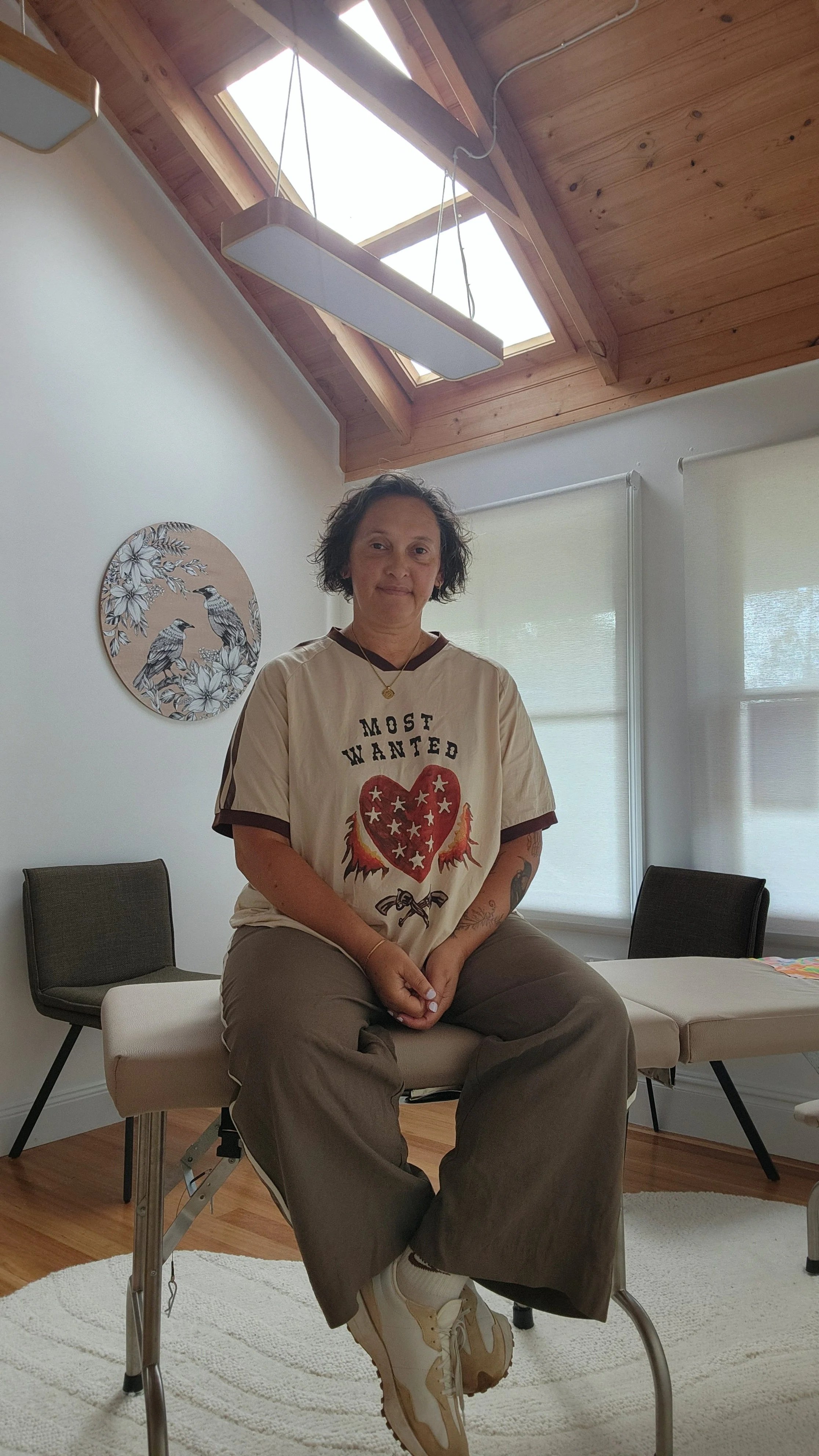 A woman sitting on an examination table in a room with wooden ceiling panels and skylights, wearing a beige shirt with a heart and 'Most Wanted' text, beige sneakers, and gray pants, with artwork of two birds on the wall behind her.