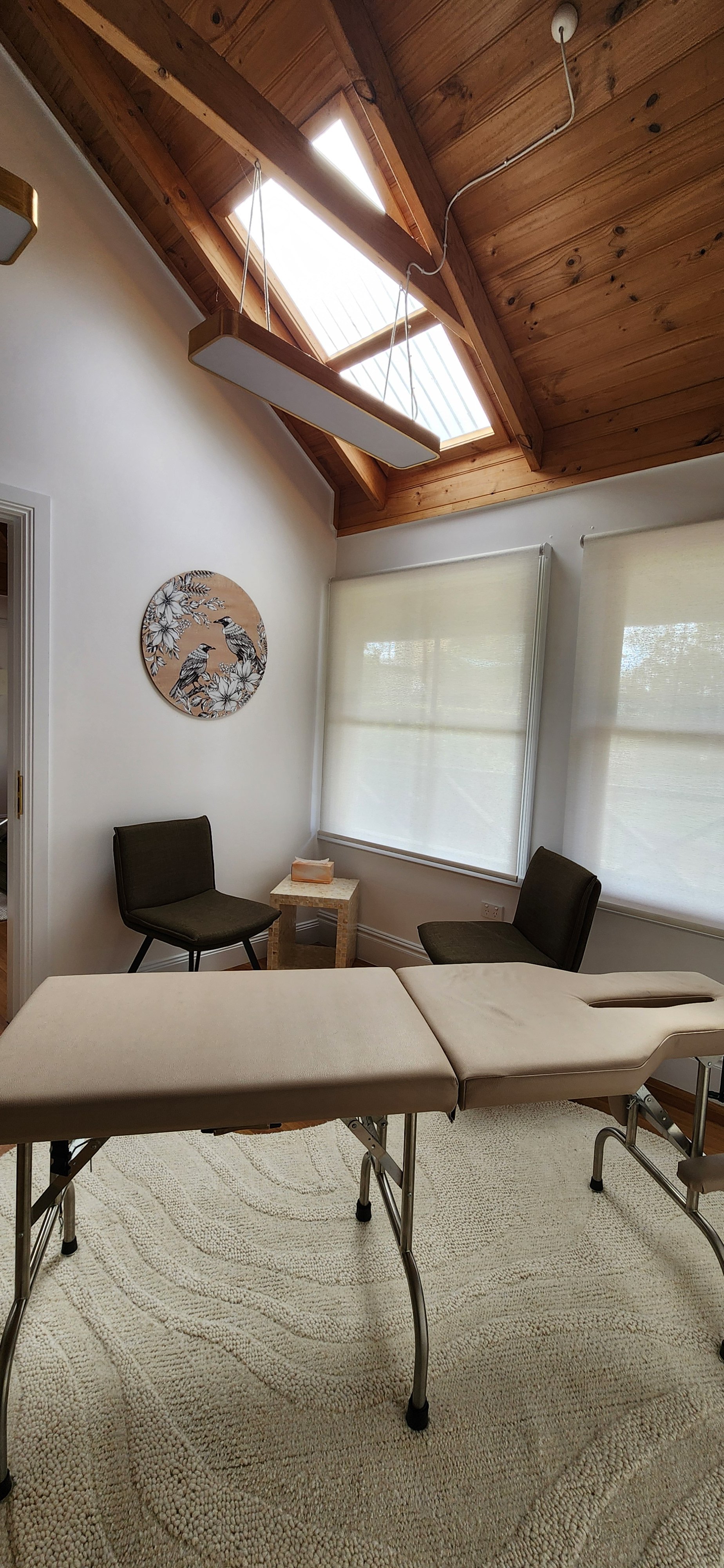 A room with a massage or examination table, two black chairs, a small side table with tissue box, sunlit windows, white walls, wooden ceiling with skylights, and a decorative circular wall art of birds.