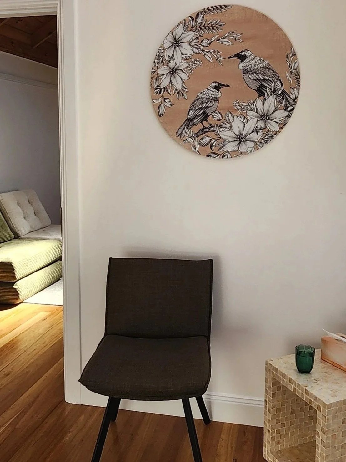 A simple room with a dark chair, a mosaic side table, and a wall hanging featuring birds and flowers.