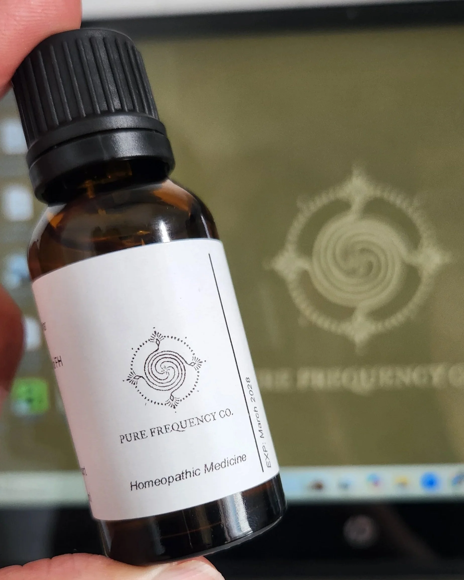 A brown glass bottle labeled 'Pure Frequency Co., Homeopathic Medicine' being held in front of a computer monitor with a logo matching the bottle's label.