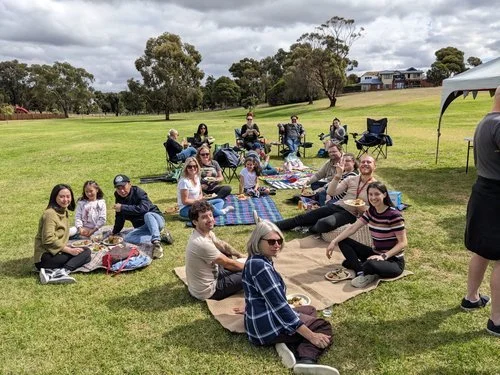 A group of people having a picnic on a grassy field with trees in the background. They are seated on blankets and folding chairs, enjoying food and drinks under a cloudy sky.