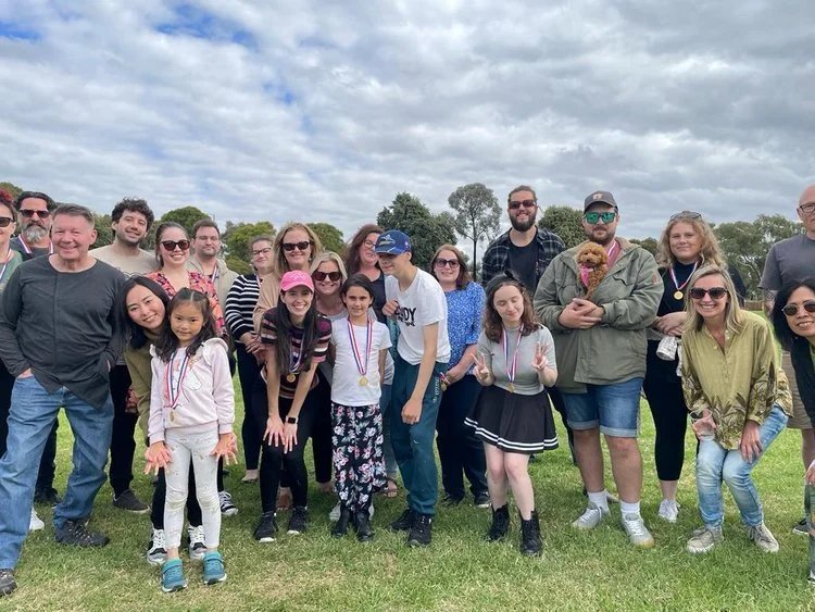 Group of people outdoors, smiling, some wearing medals, on a grassy field under a cloudy sky.
