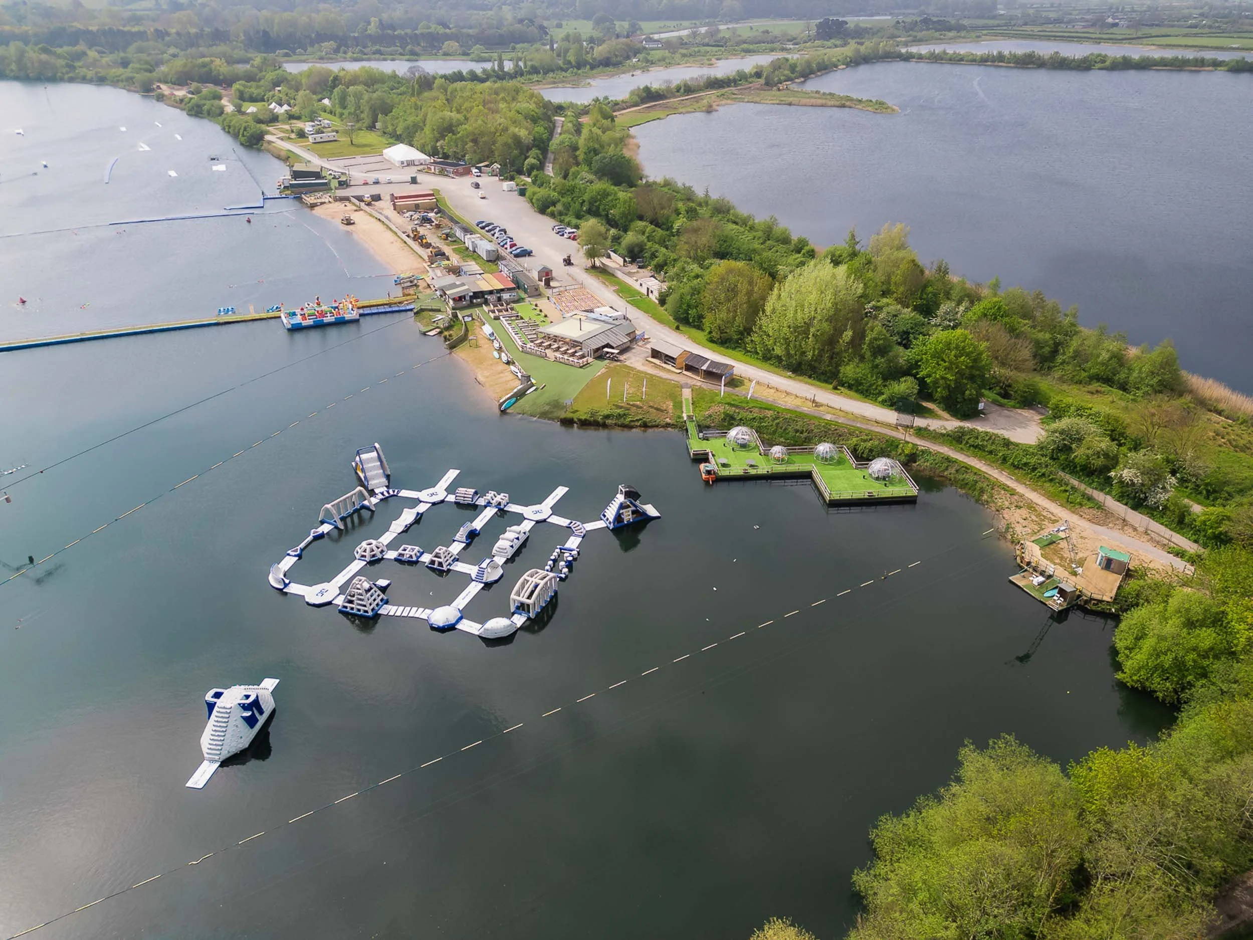 Aerial view of a water sports facility on a lake, featuring a large inflatable obstacle course surrounded by green trees in the background.