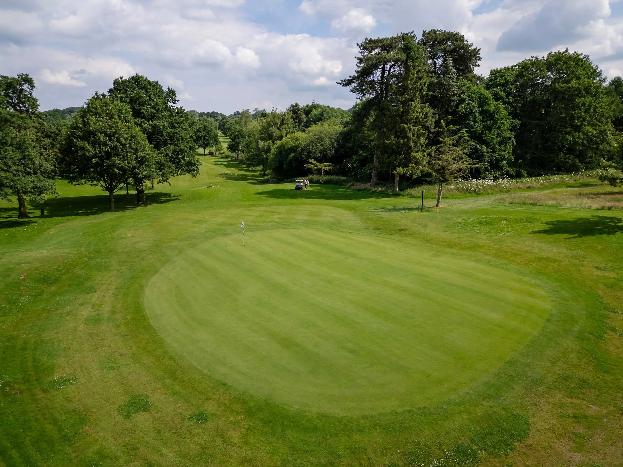 Aerial view of a golf green and fairway stretching into the distance, bordered by lush green trees under a blue sky with clouds.