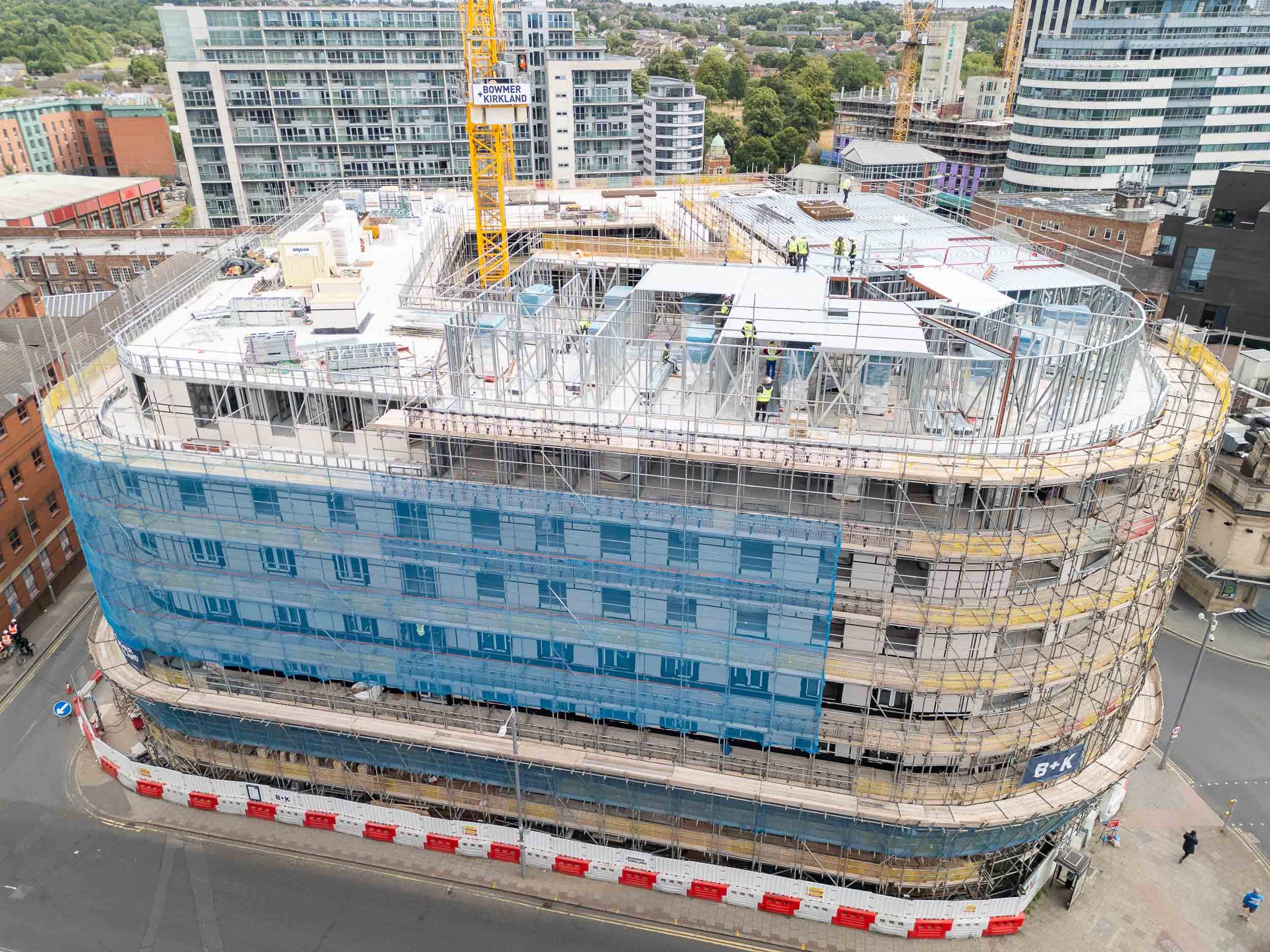 Aerial drone view of a partially constructed apartment building in a busy city centre, surrounded by roads and neighbouring buildings.