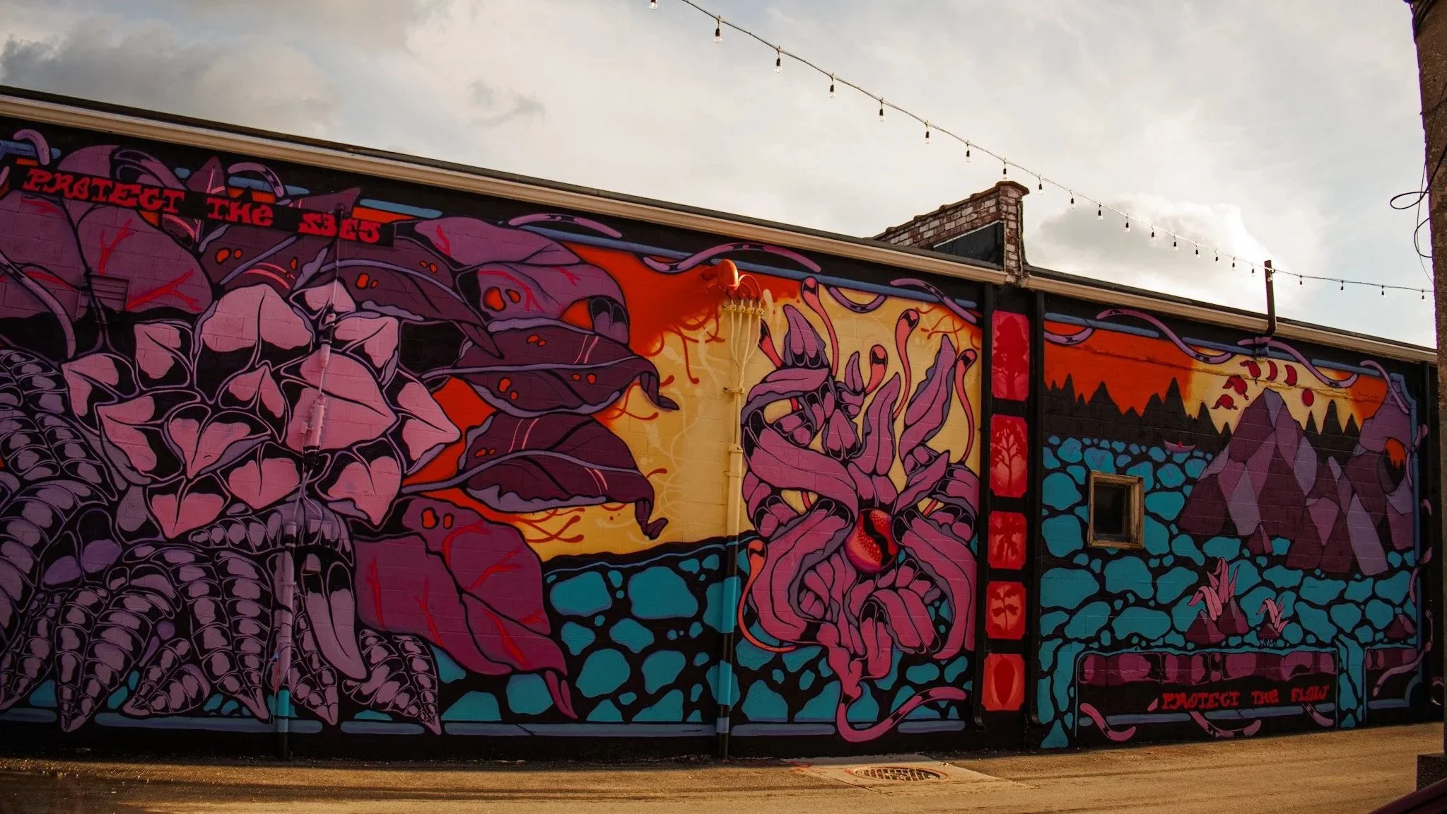 Mural colorido com desenhos abstratos de cores roxa, laranja, azul e preta em uma parede de fundo preto, com luzes penduradas acima