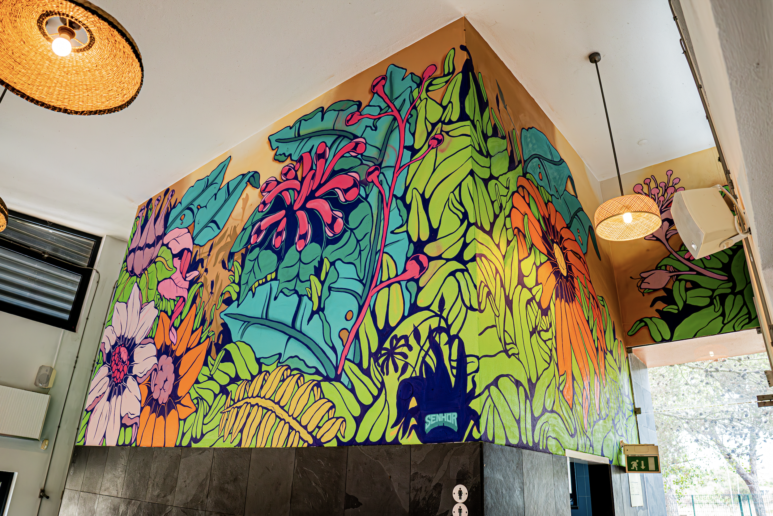 Mural colorido de flores e folhas em um espaço interno, iluminado por luminária de tecido trançado, com teto branco e parede de tijolos à vista no canto direito.