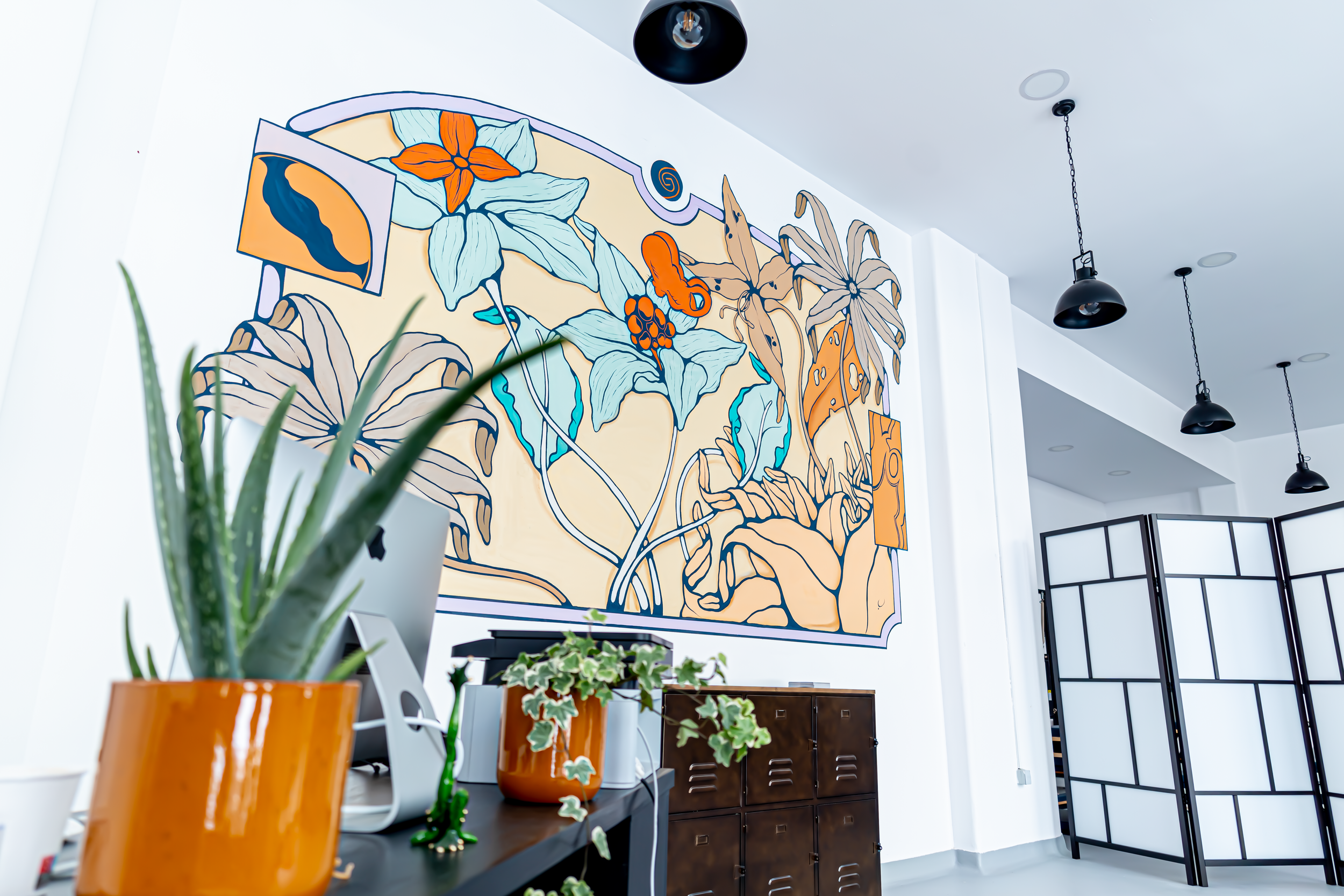 Relevo de parede com ilustração de flores, pássaros e insetos, decorando o interior de um espaço moderno com luminárias pretas suspensas e plantas em vasos de cerâmica laranja na frente.