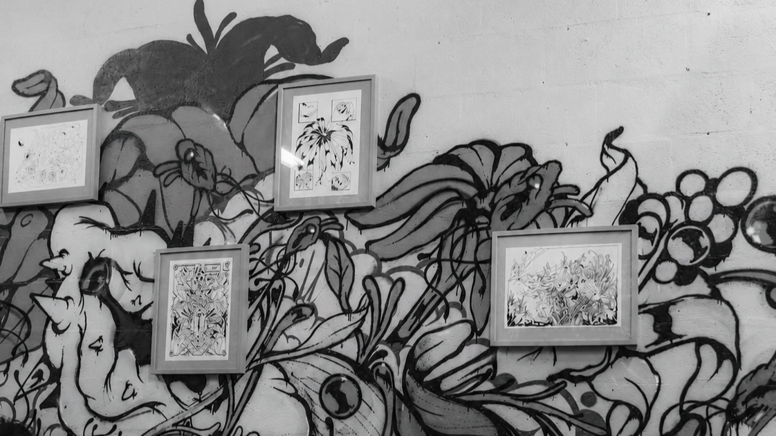 Imagem de uma parede com grafite em preto e branco com várias plantas e flores, decorada com quatro molduras com desenhos artísticos.