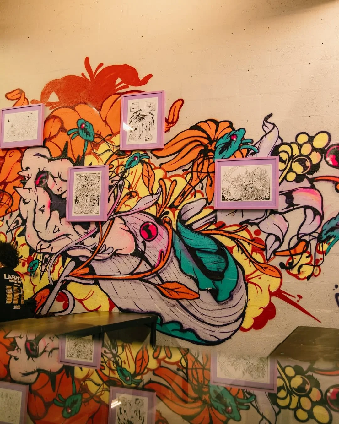 Mural com desenhos em estilo de arte urbana com várias cores vibrantes, incluindo laranja, verde, preto, roxo e amarelo. Existem quadros de desenhos emoldurados na parede, que parecem ser desenhos em preto e branco.