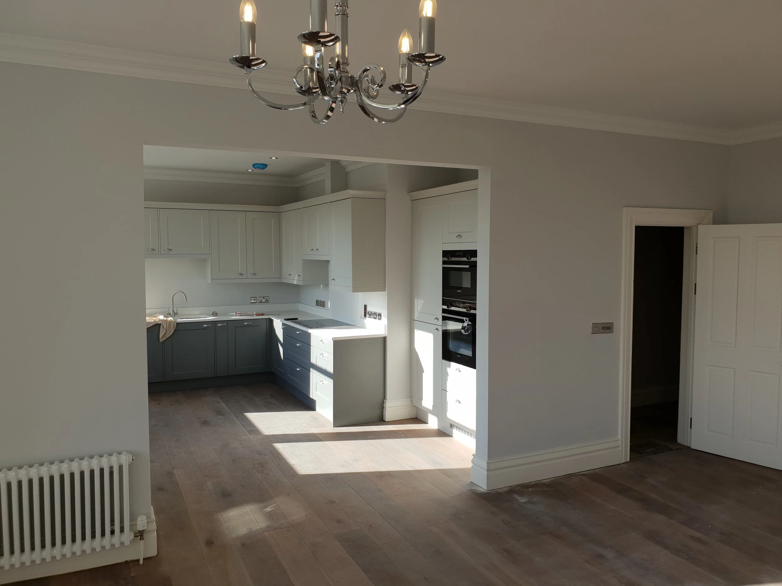 Modern kitchen with white cabinets, built-in oven, gray lower cabinets, and a chandelier hanging from the ceiling. Natural light illuminates the space, and there is a radiator in the foreground.