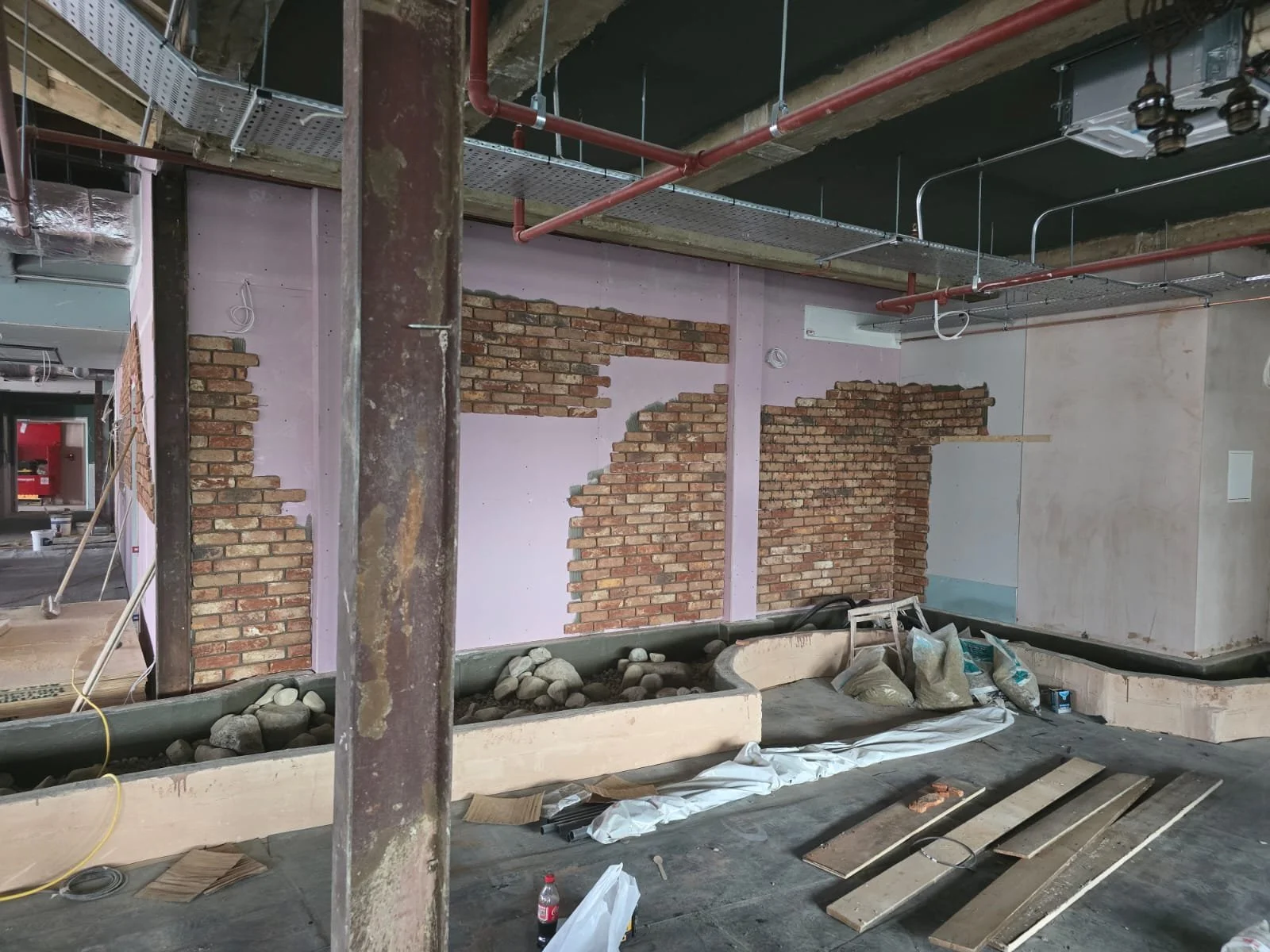 Interior of a building under construction with exposed brick walls, construction materials, and pipes on the ceiling.