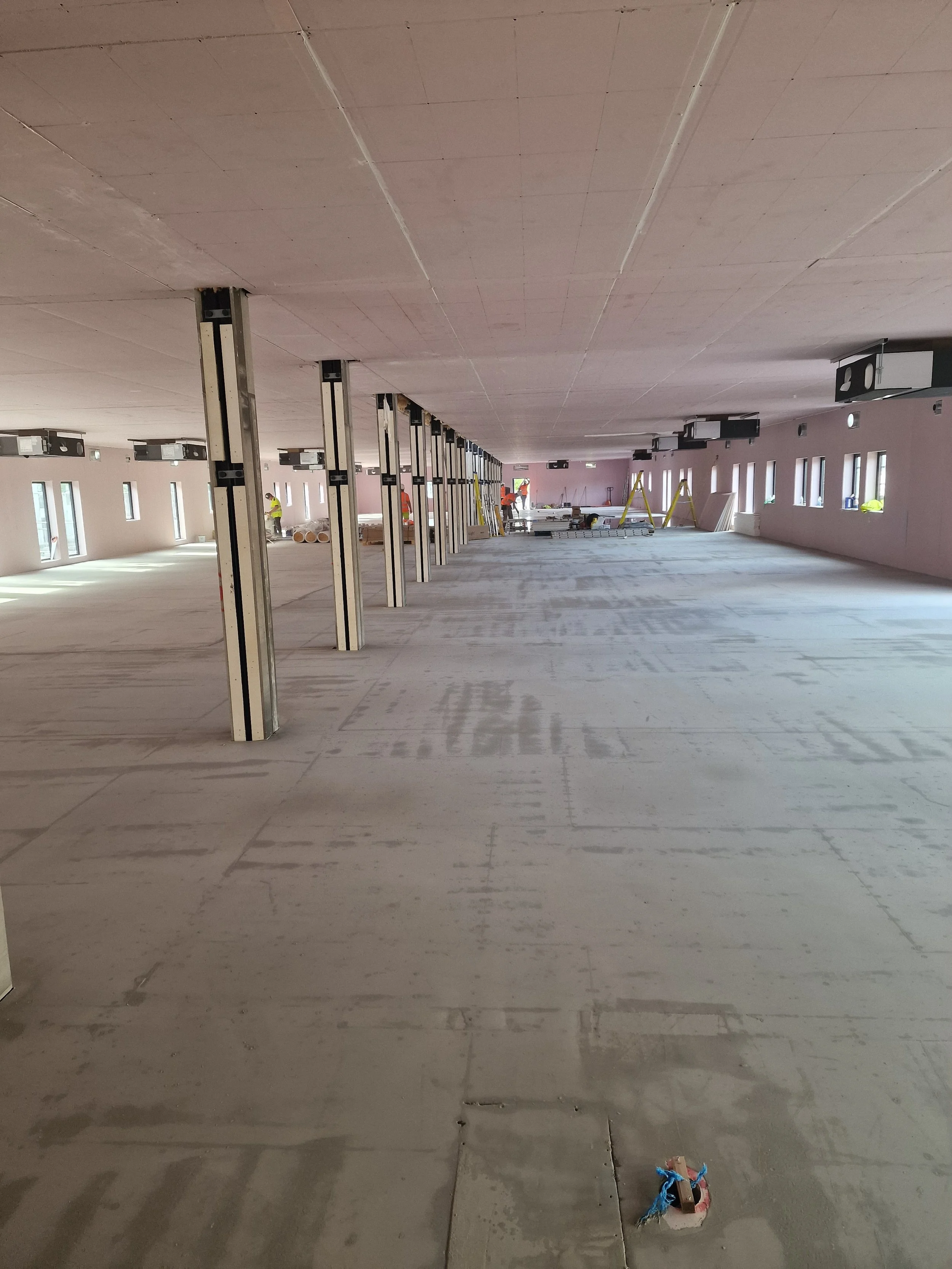 Empty room under construction with exposed support beams, construction workers, and ladders in the background.