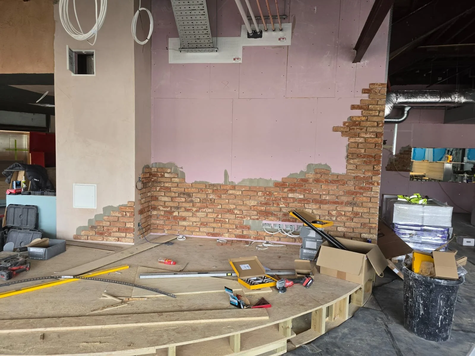 Interior of a construction site where wall partially torn down revealing brickwork behind drywall, construction tools, and materials scattered on a curved wooden platform.