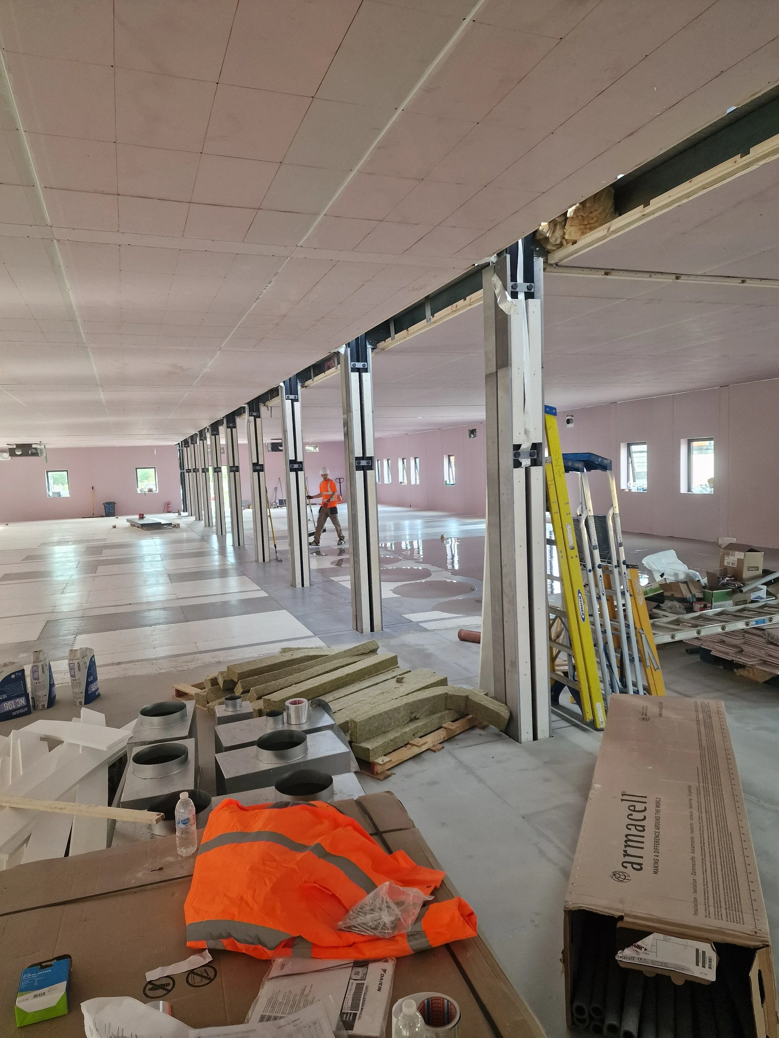 Inside a building under construction with metal support beams, construction materials, and a worker wearing an orange safety vest and hard hat.