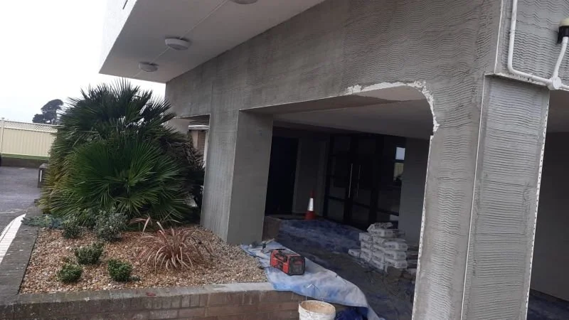 Construction site with unfinished building exterior, construction materials, and a small landscaped area with plants and rocks.