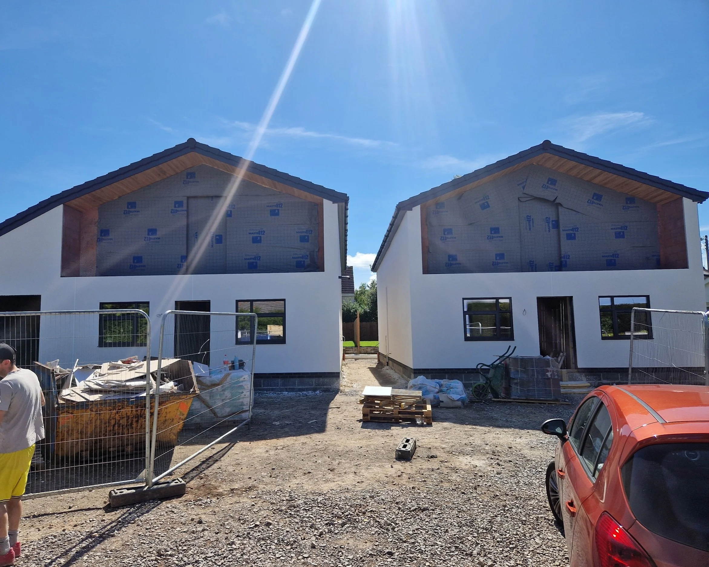Construction site with two partially built houses covered in insulation material, construction fencing, construction debris, and a red car parked in front.