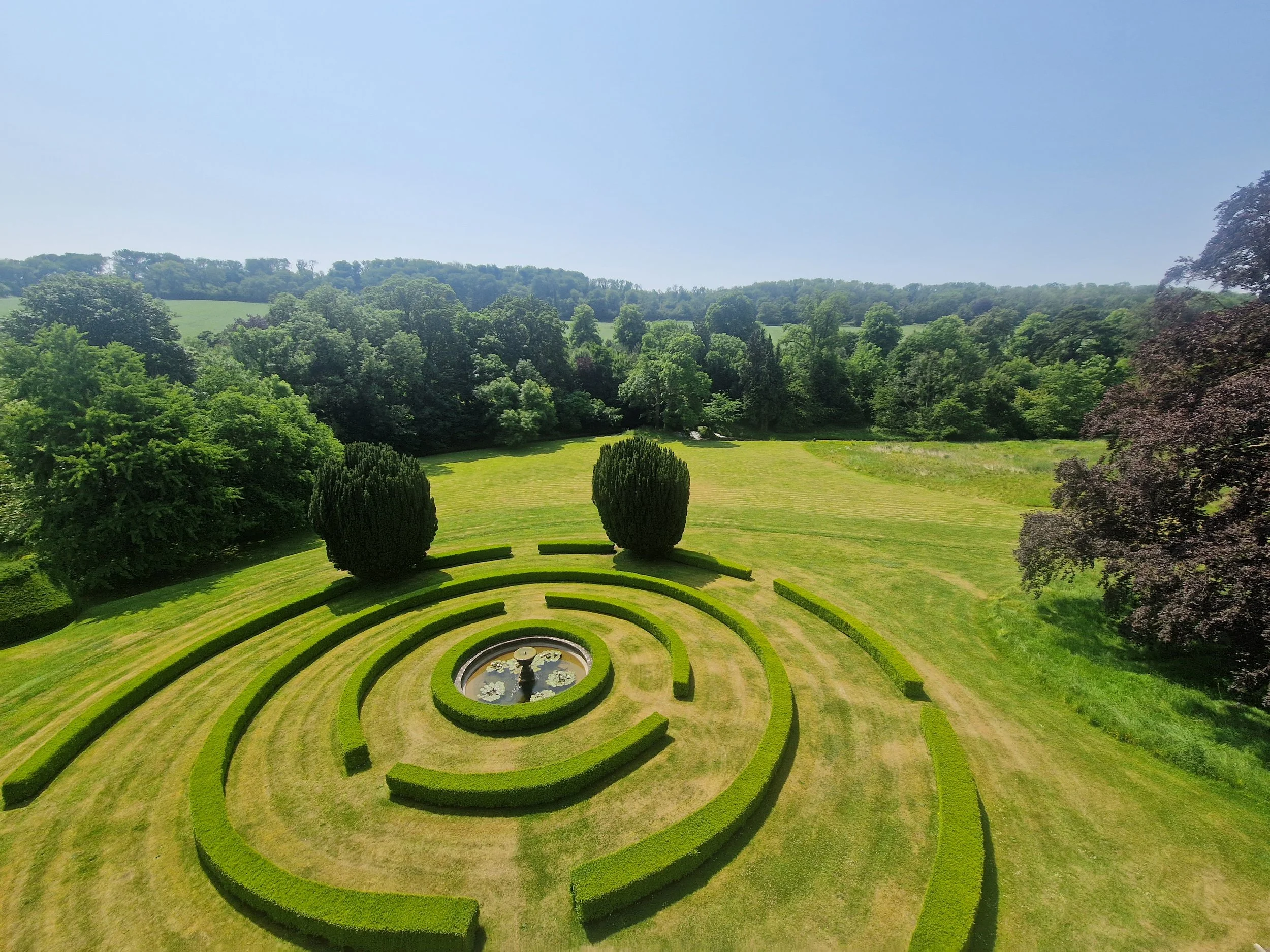 A circular garden with neatly trimmed hedges arranged in concentric circles around a fountain with lily pads, set against a backdrop of lush green trees and a clear blue sky.