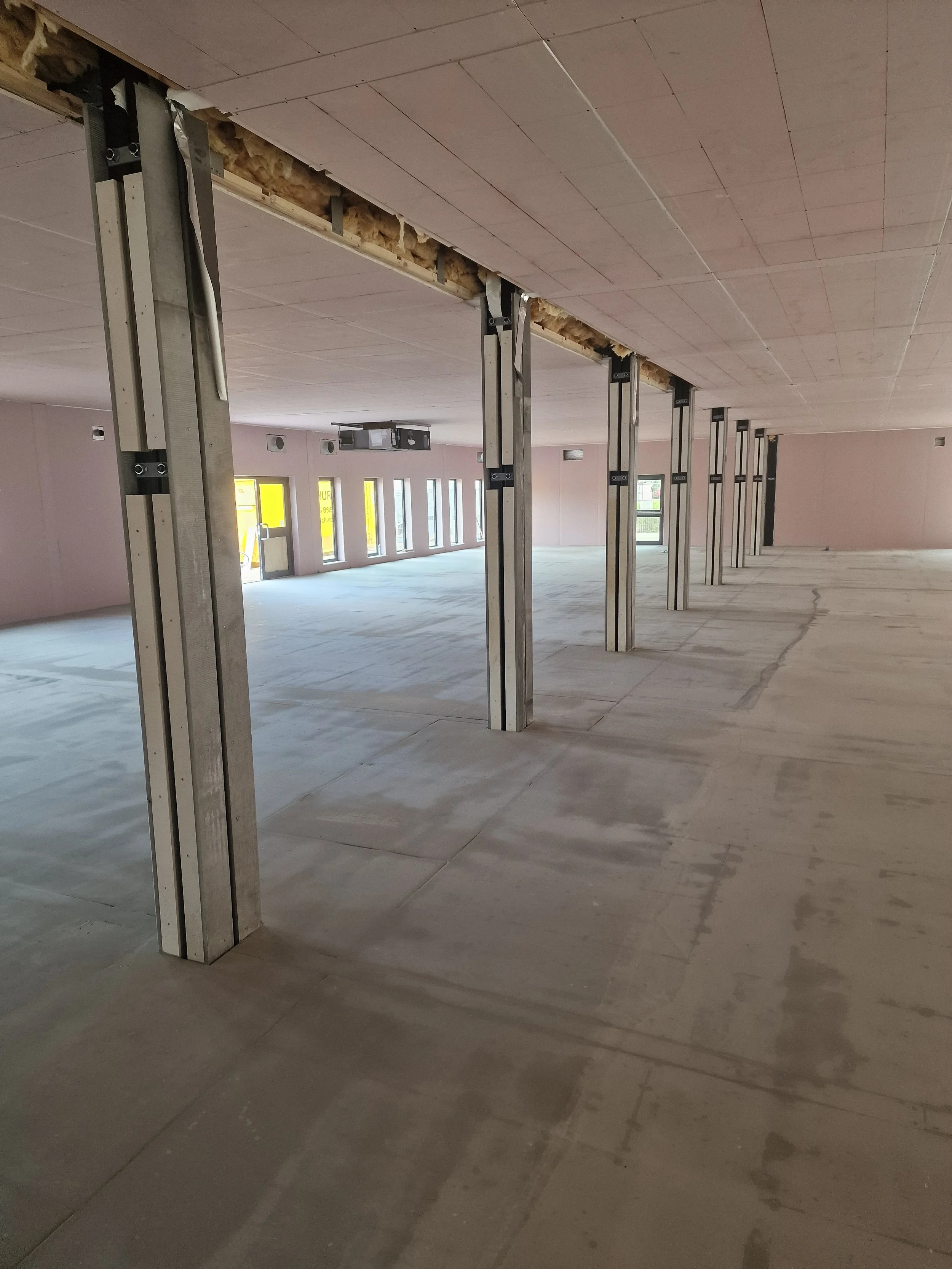 Interior of a building under construction with exposed metal studs and unfinished ceiling, large open floor space, and natural light coming through multiple windows.