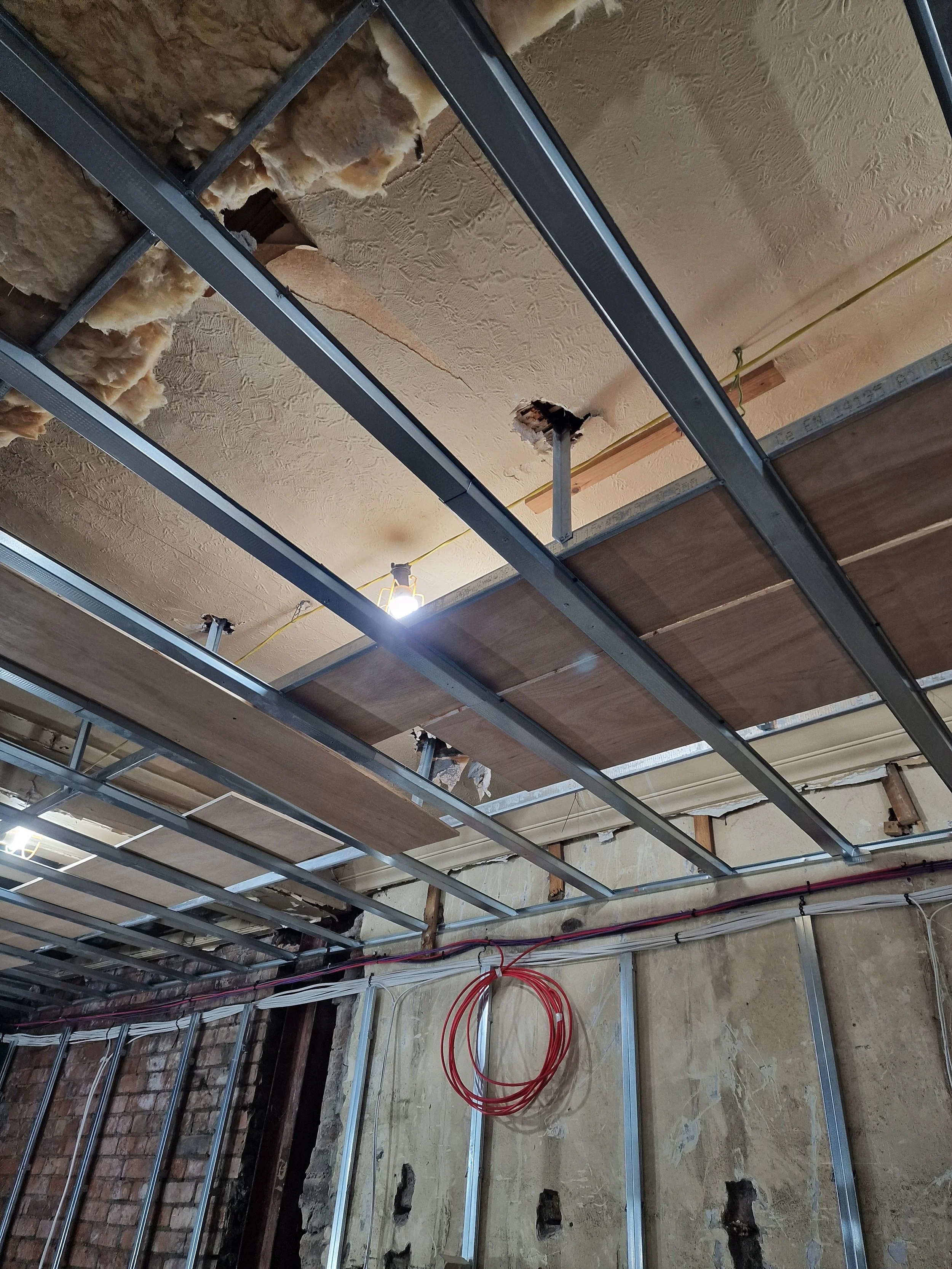 Construction framing in progress with metal studs and exposed insulation on the ceiling. Red electrical cables are coiled on the unfinished floor, and pipes are visible along the wall.