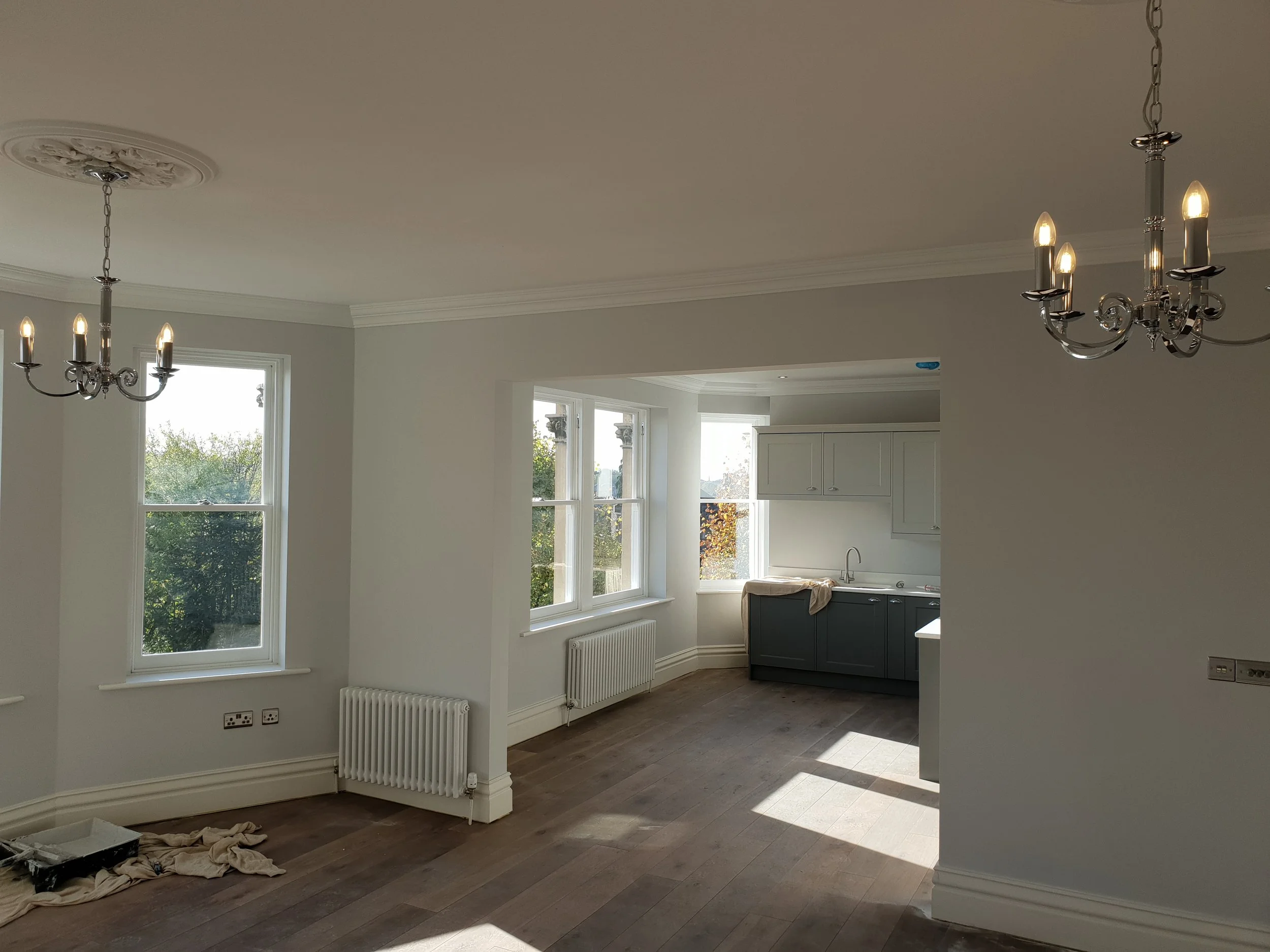 Unfinished interior of a house with large windows, a chandelier, white walls, and a kitchen area in the background. Construction materials and tools are on the floor.