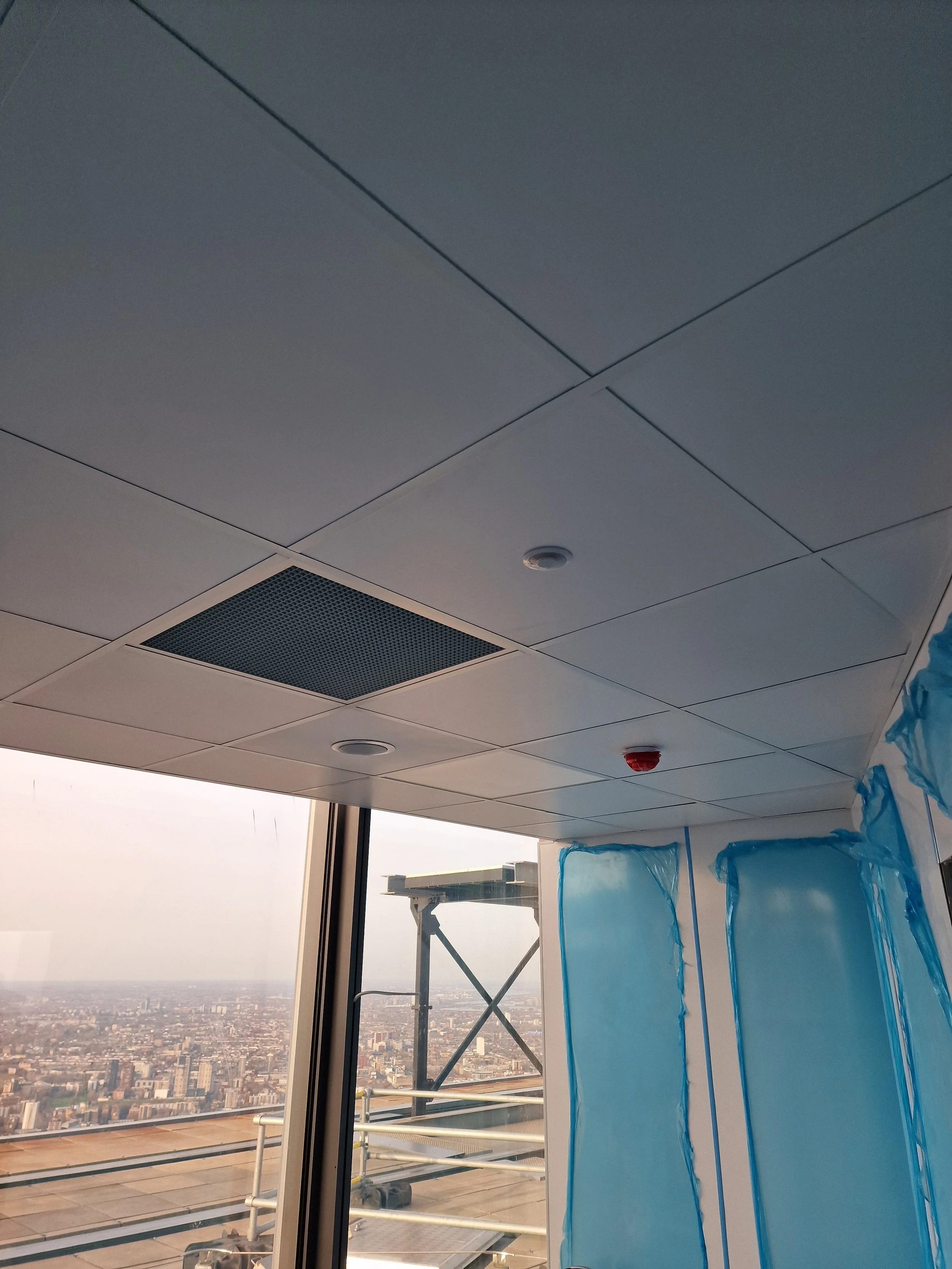 Interior view of a high-rise building with a partial view of a cityscape outside the window, under construction scaffolding, with some blue plastic sheets covering part of the interior ceiling.