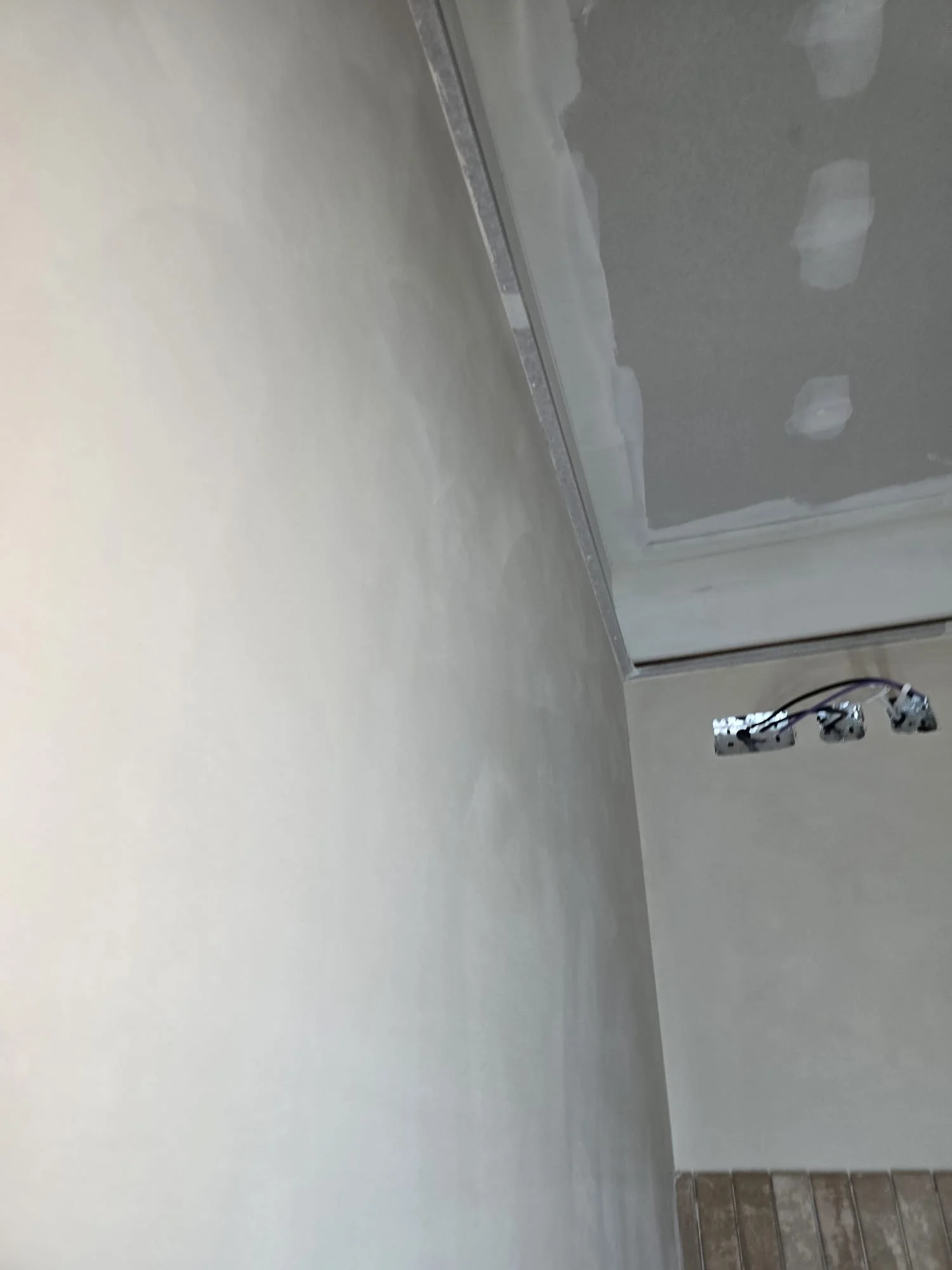 Interior wall with drywall and a metal trim, electrical wires hanging near the bottom, and a small section of tiled floor in the lower right corner.