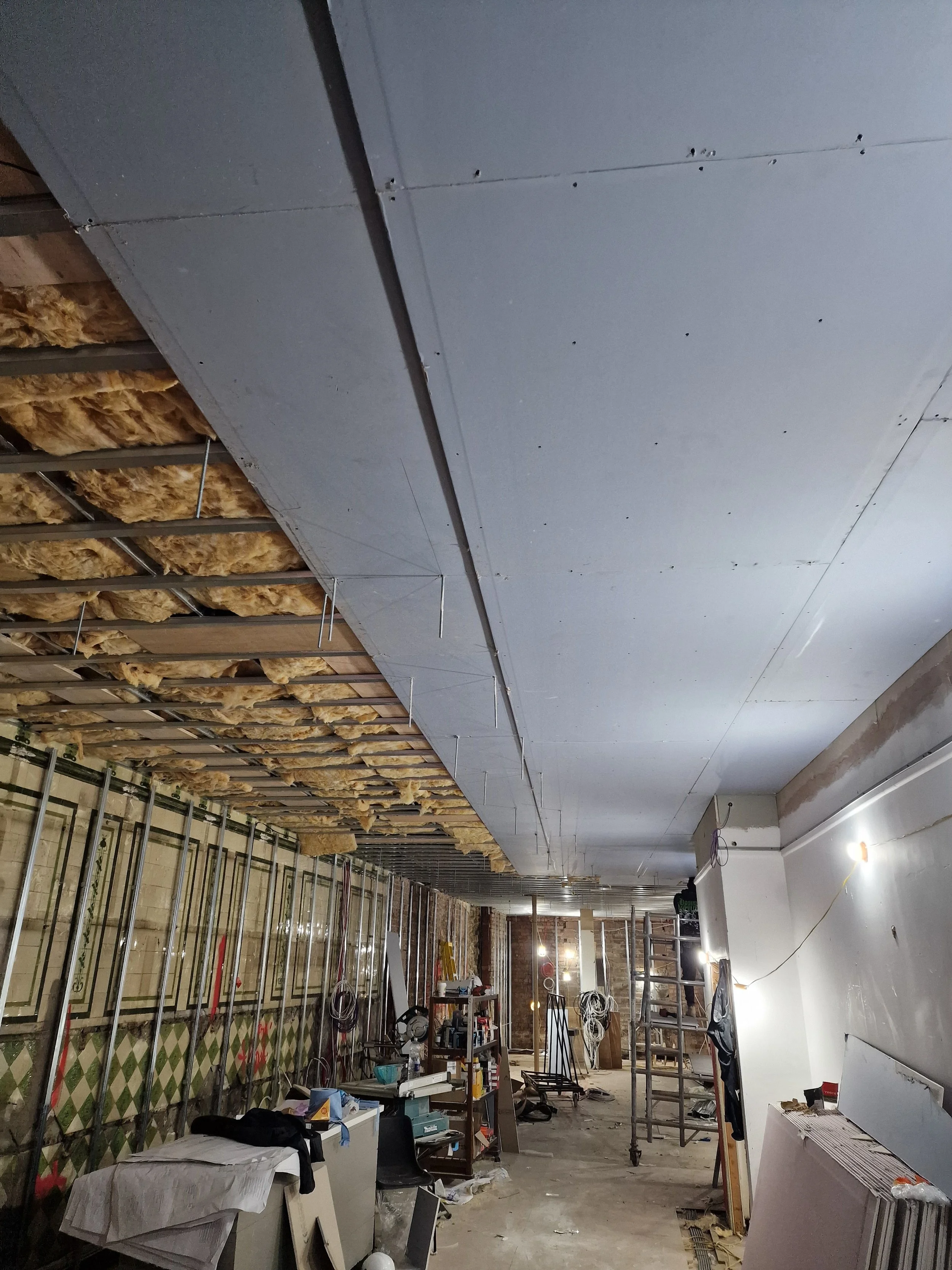 A construction site inside a building where drywall is being installed on the ceiling, with some insulation visible in the upper left, and various tools, furniture, and construction materials scattered around the unfinished room.