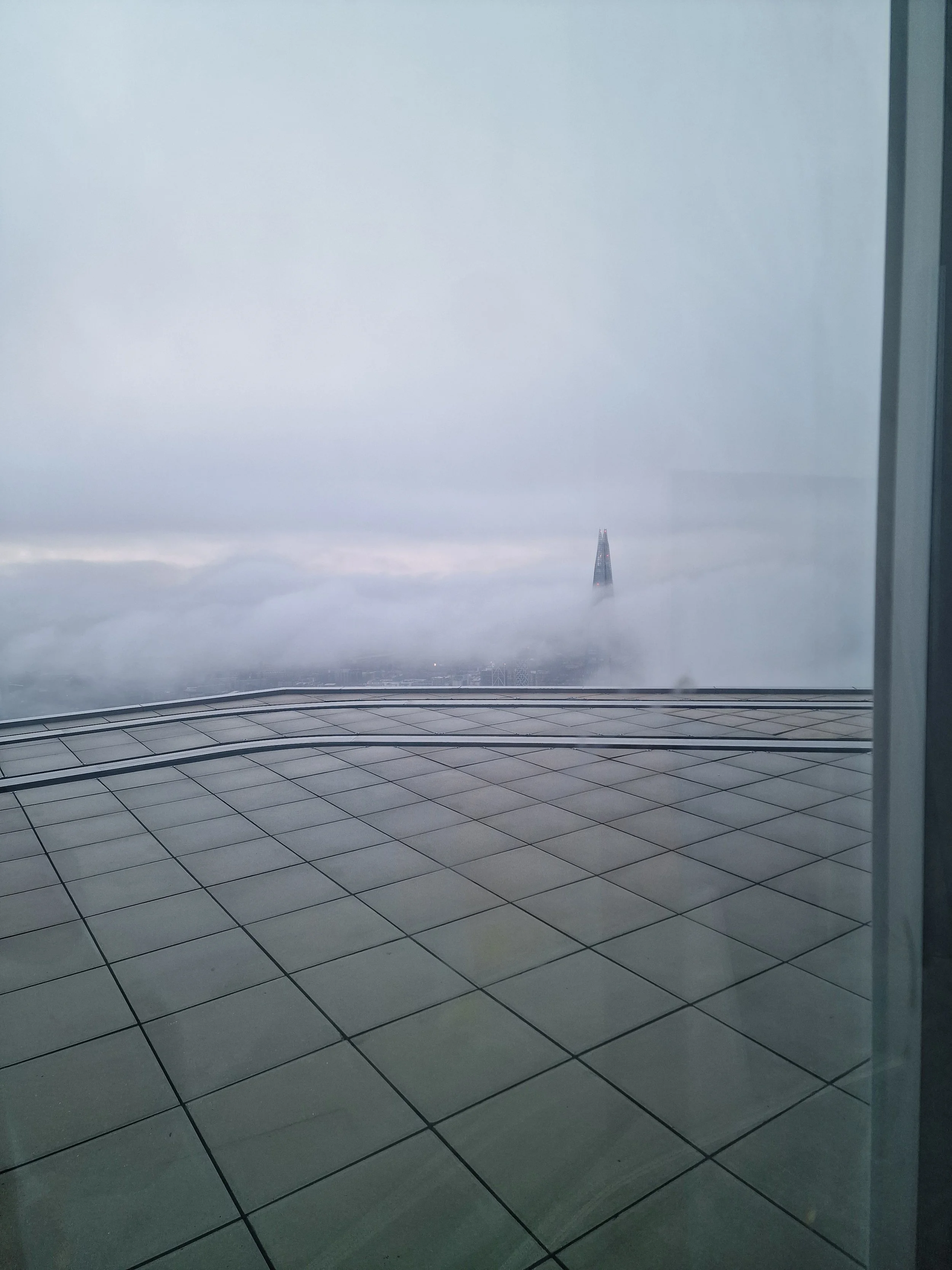 View of a tall building emerging through a thick layer of clouds, photographed from inside a building with tiled flooring.