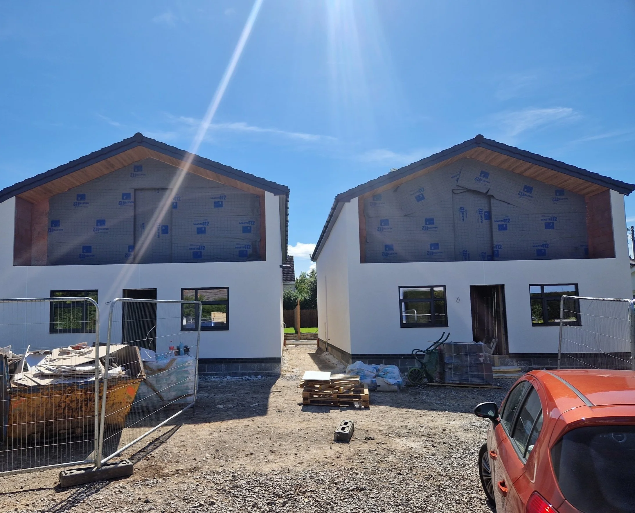 Two houses under construction with partially finished exteriors, construction materials and equipment in the foreground, and a clear blue sky with bright sunlight.