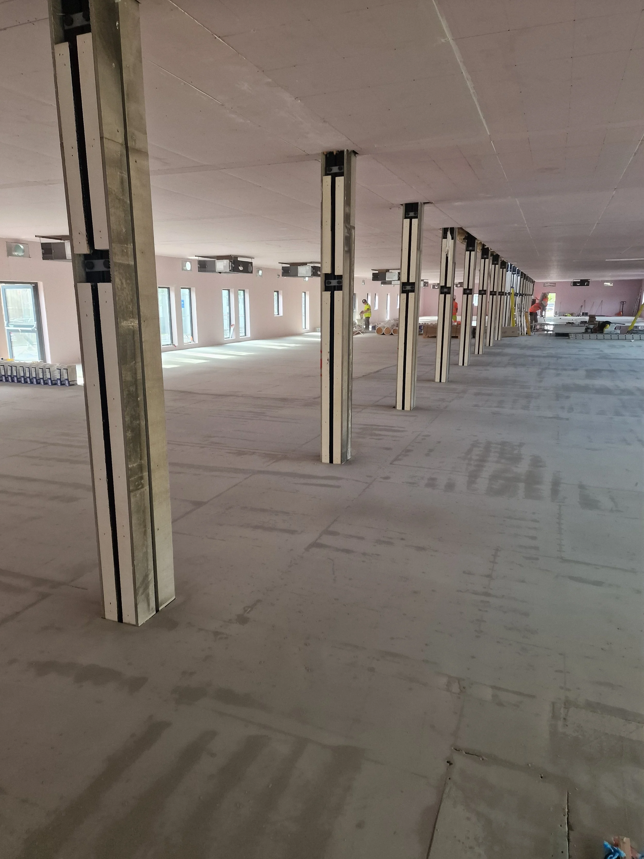 Construction site with steel poles, workers in safety vests, and ladder, in an open room with windows and unfinished walls.