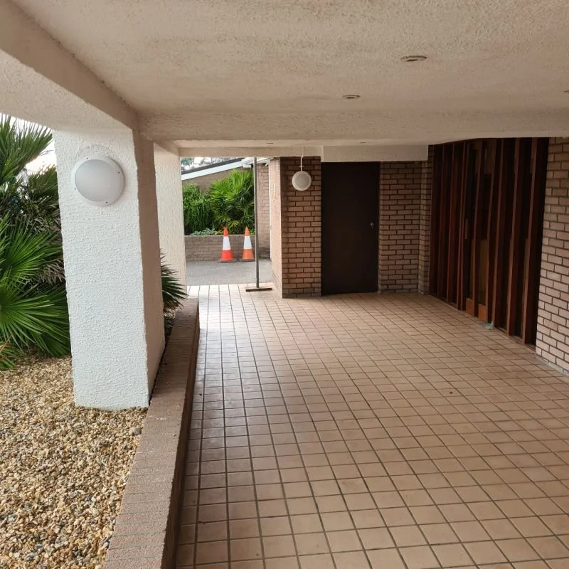 Empty covered porch area with tiled flooring, brick walls, and hanging light fixtures, leading to a sidewalk with two orange traffic cones in the background.