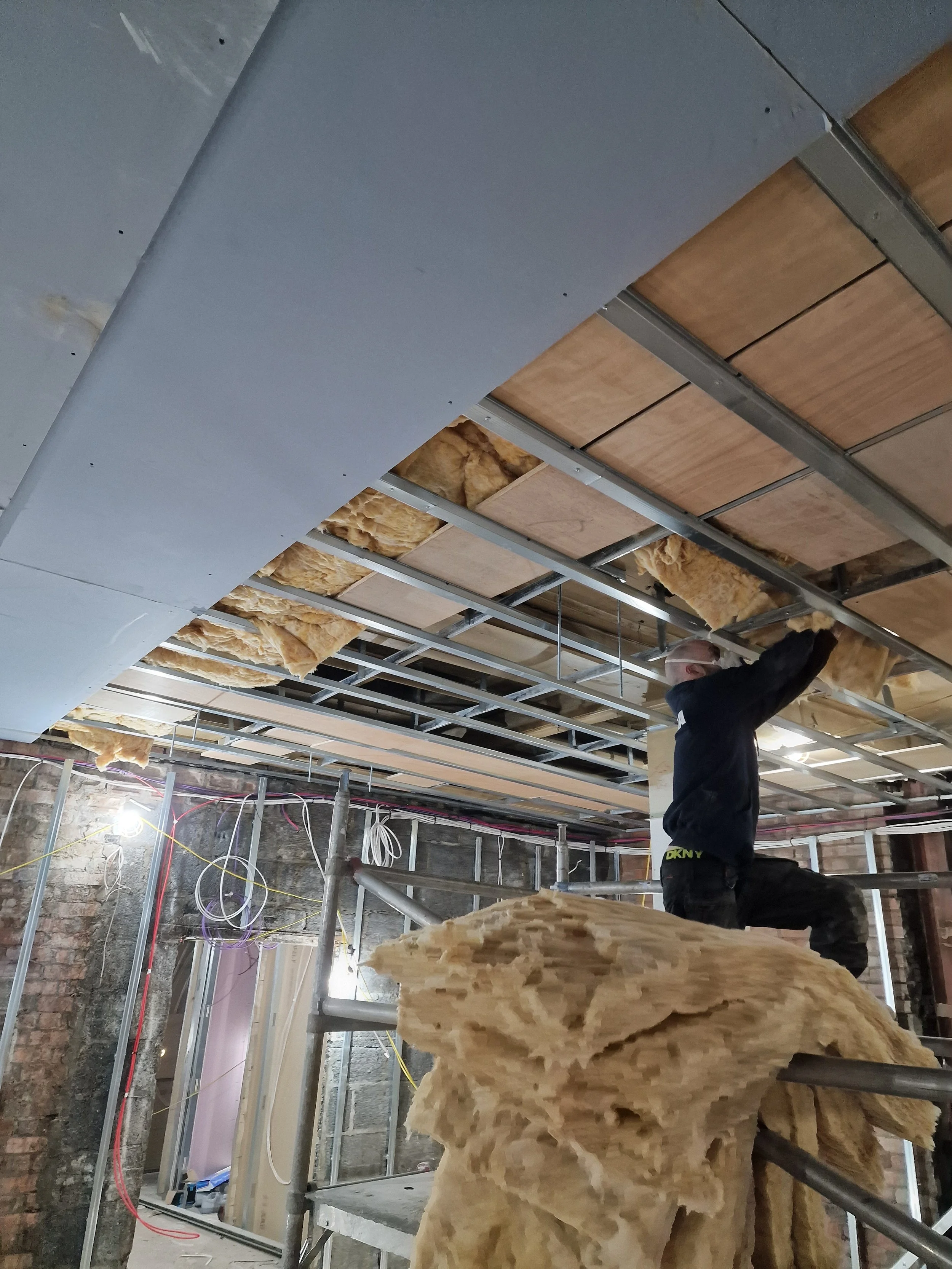 A construction worker installing drywall on a ceiling framework with insulation visible. The worker is standing on scaffolding and working with tools, in an unfinished building interior with exposed brick walls and electrical wiring.