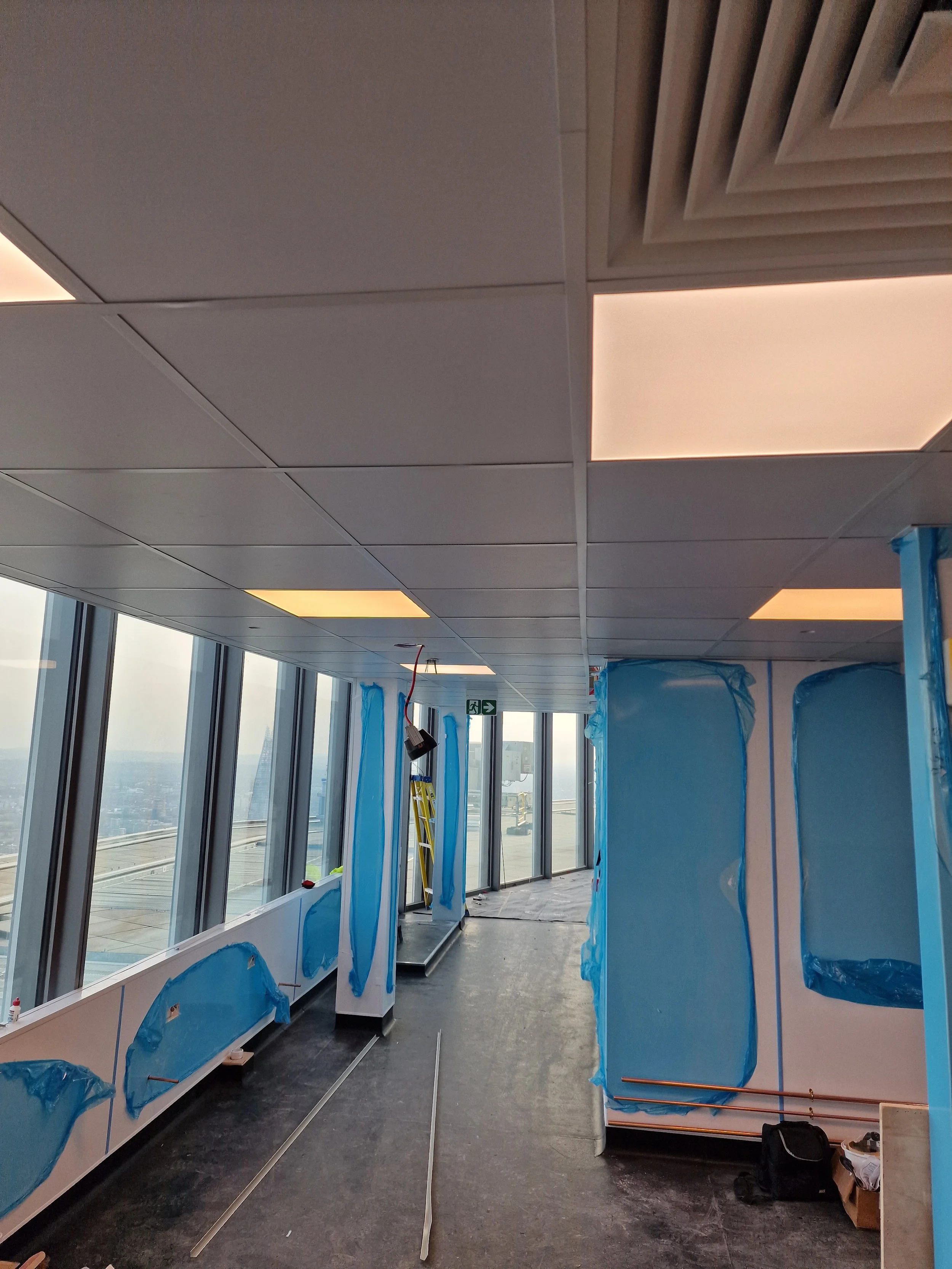 Interior of a building under construction with large windows, ceiling panels, and construction materials, including blue protective coverings on walls and fixtures.