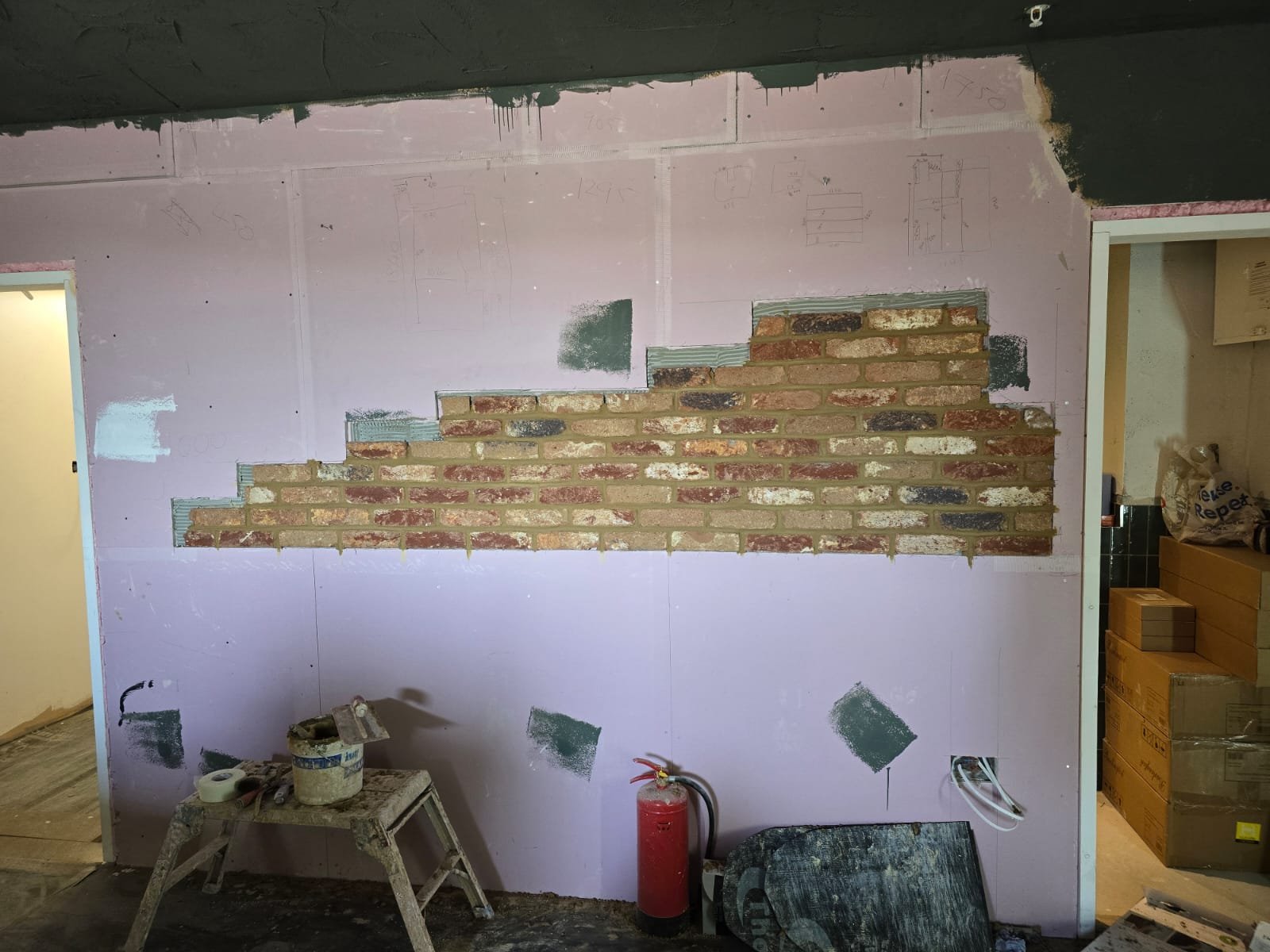 Interior wall under construction with a partial brick wall section and pink drywall panels marked with construction sketches, with tools, a fire extinguisher, and construction materials on the floor.