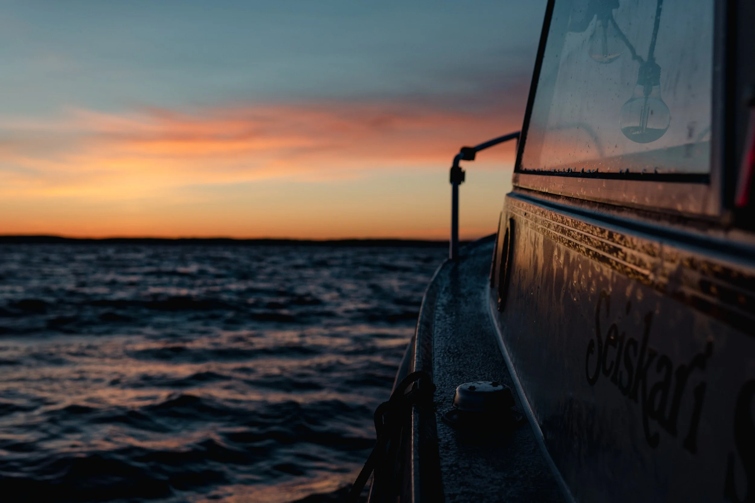 Auringonlasku hehkuu taivaanrannan yllä, nähtävissä osa venekannen sivusta ja perävaijereja, jotka roikkuvat veden päällä.