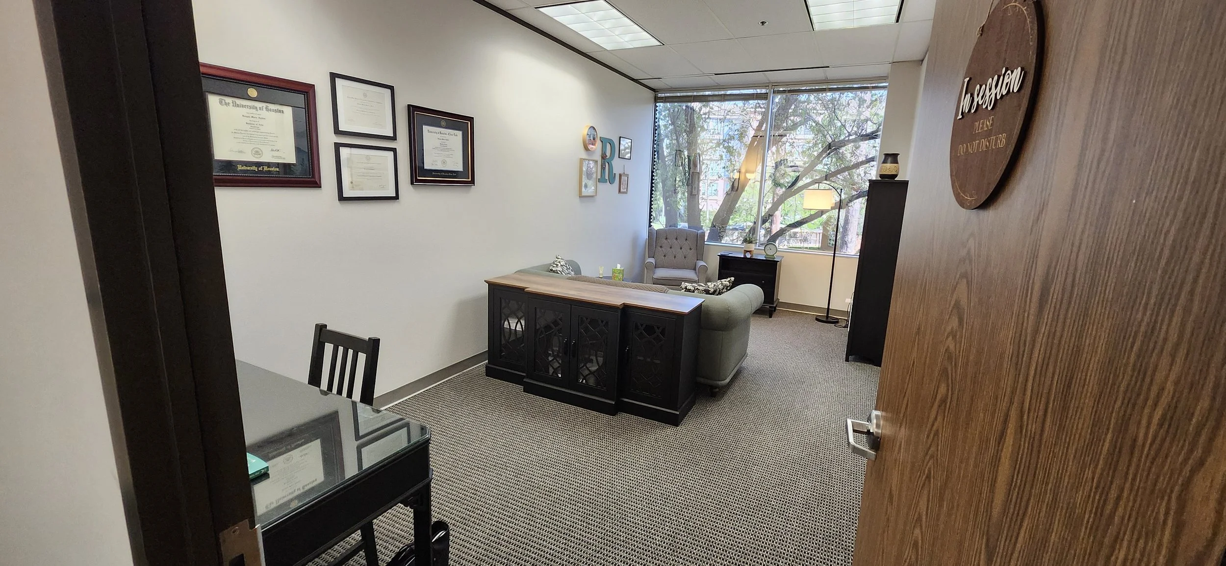 View of a professional office from an open wooden door, featuring a seating area with a sofa, armchair, and a side table, decorated with framed diplomas and artwork on the walls, large window with trees outside, and various decorative items.