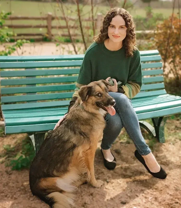 A woman with curly brown hair wearing a green sweater and jeans sitting on a park bench with a medium-sized brown and black dog with its tongue out.