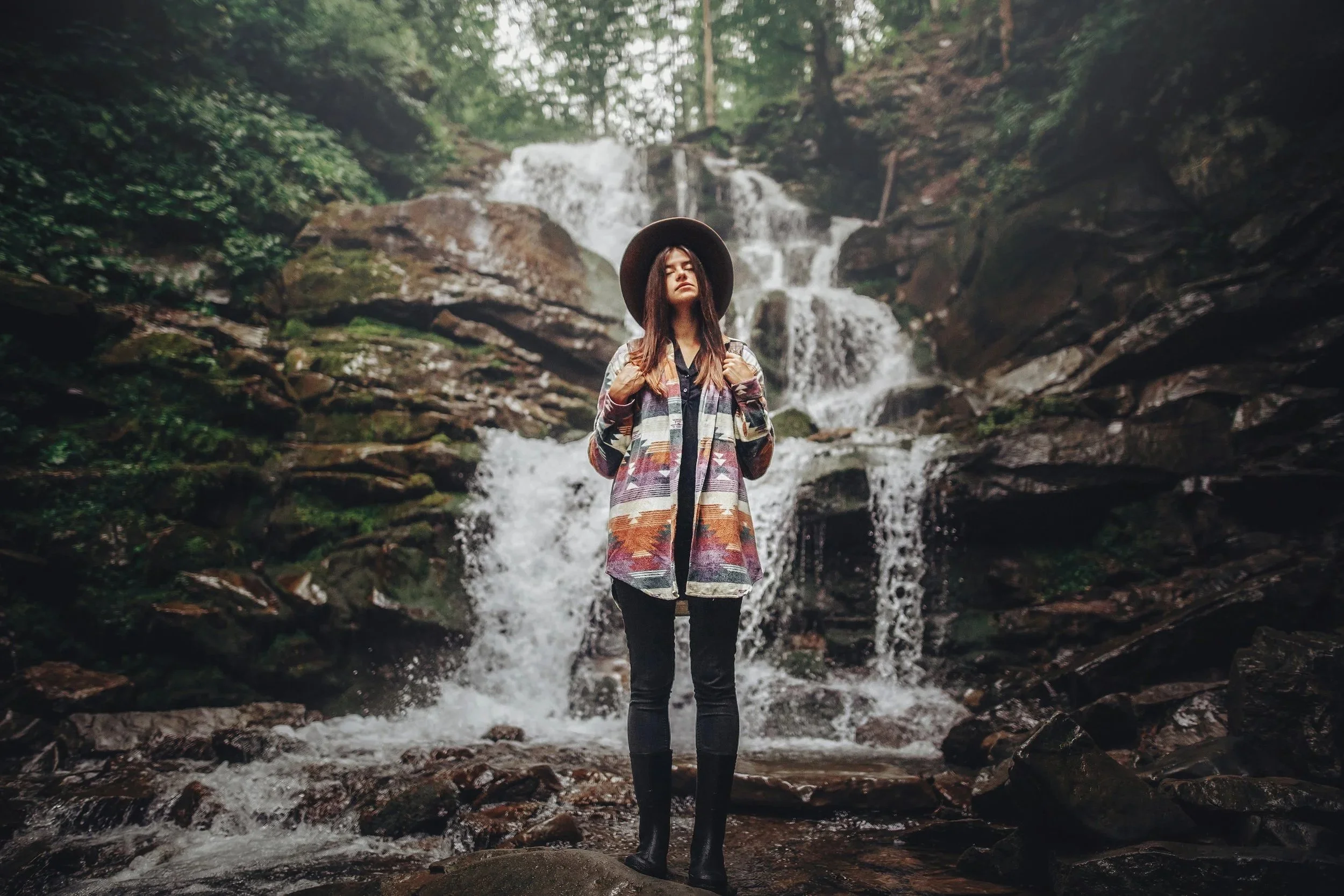 A woman standing in front of a waterfall and rocks in a forest, wearing a hat, a colorful patterned coat, and black pants.