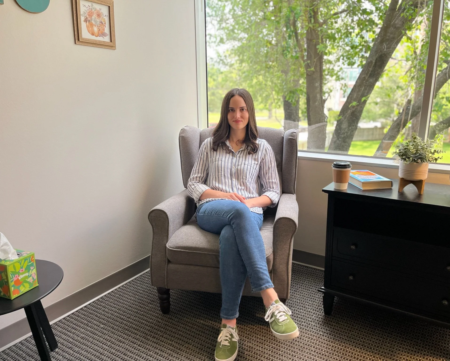 A woman sitting in a gray armchair in an office, next to a window with green trees outside. She is wearing a striped button-up shirt, jeans, and green sneakers. A table beside her has a disposable coffee cup, a book, and a plant, with a colorful box on another table in the foreground.