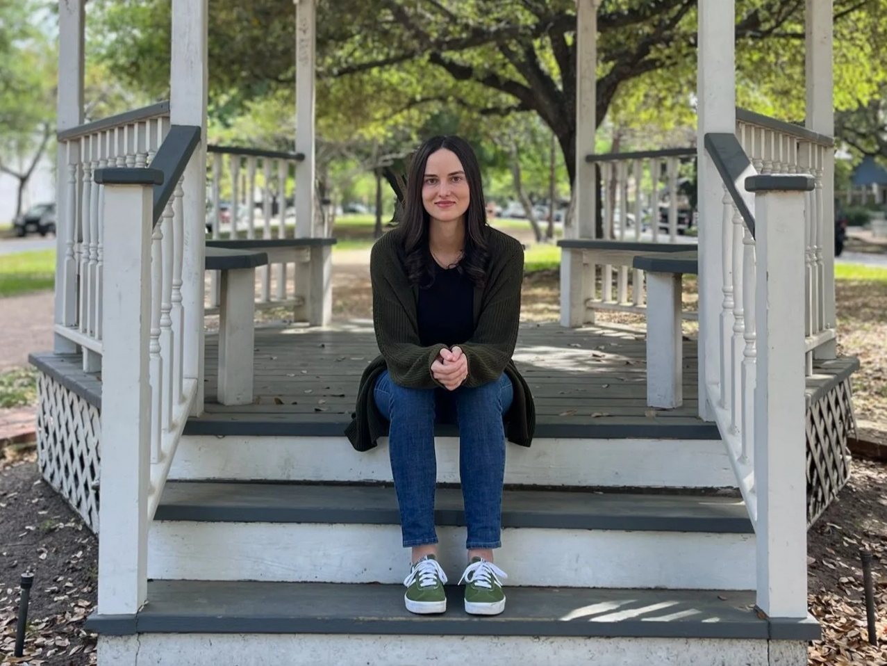 A young woman with long black hair, sitting on the steps of a white wooden gazebo in a park with green trees and grass in the background.