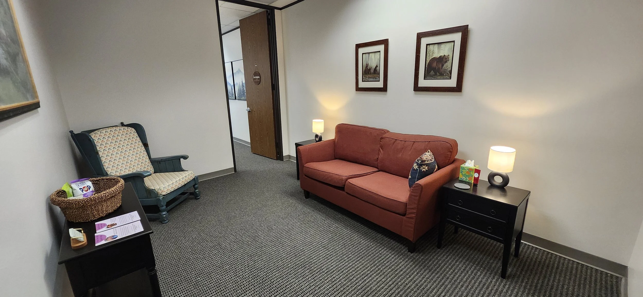 A cozy waiting room with a red sofa, a black side table with a lamp, a patterned armchair, and a small table with snacks and pamphlets. Two framed pictures of bears and a forest scene hang on the wall, and there is an open door leading to a hallway w