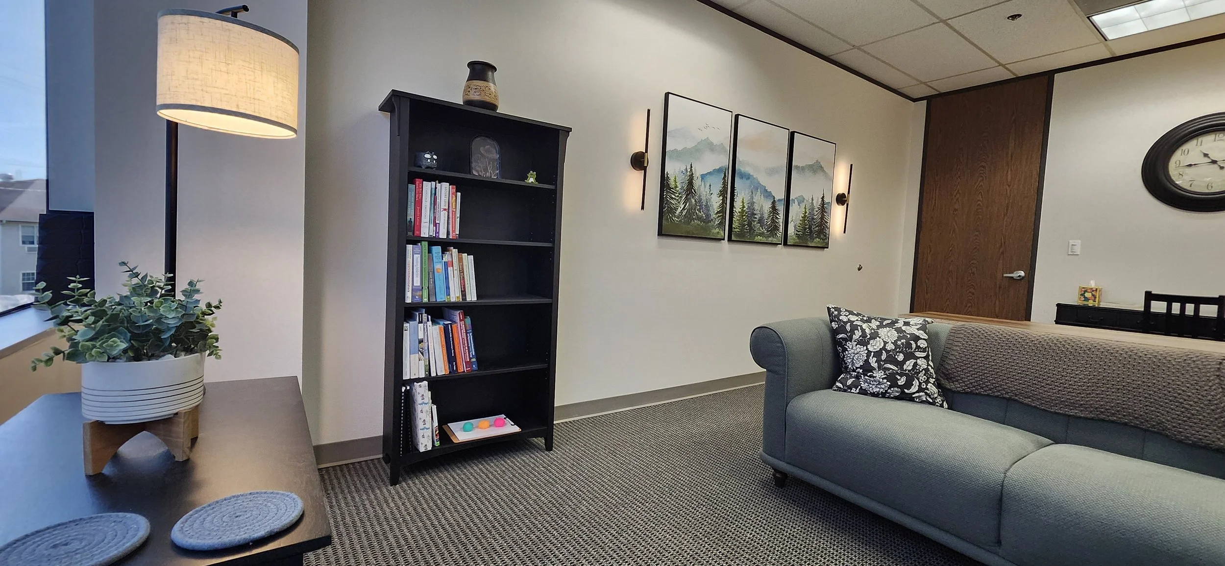 Office room with a window, a beige sofa with a floral pillow, a black bookshelf filled with books, a framed mountain landscape on the wall, a wall clock, and a potted plant on a table.