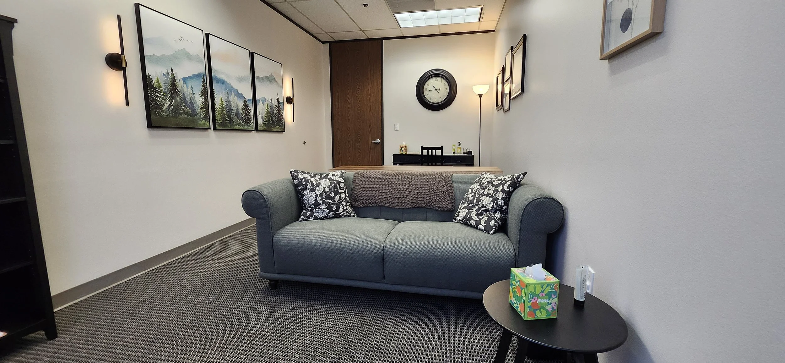 A cozy living room corner with a gray sofa, decorated with black and white floral pillows and a brown throw blanket. Above the sofa, three landscape paintings hang on the wall, illuminated by wall sconces. In the background, a wall clock, a floor lam