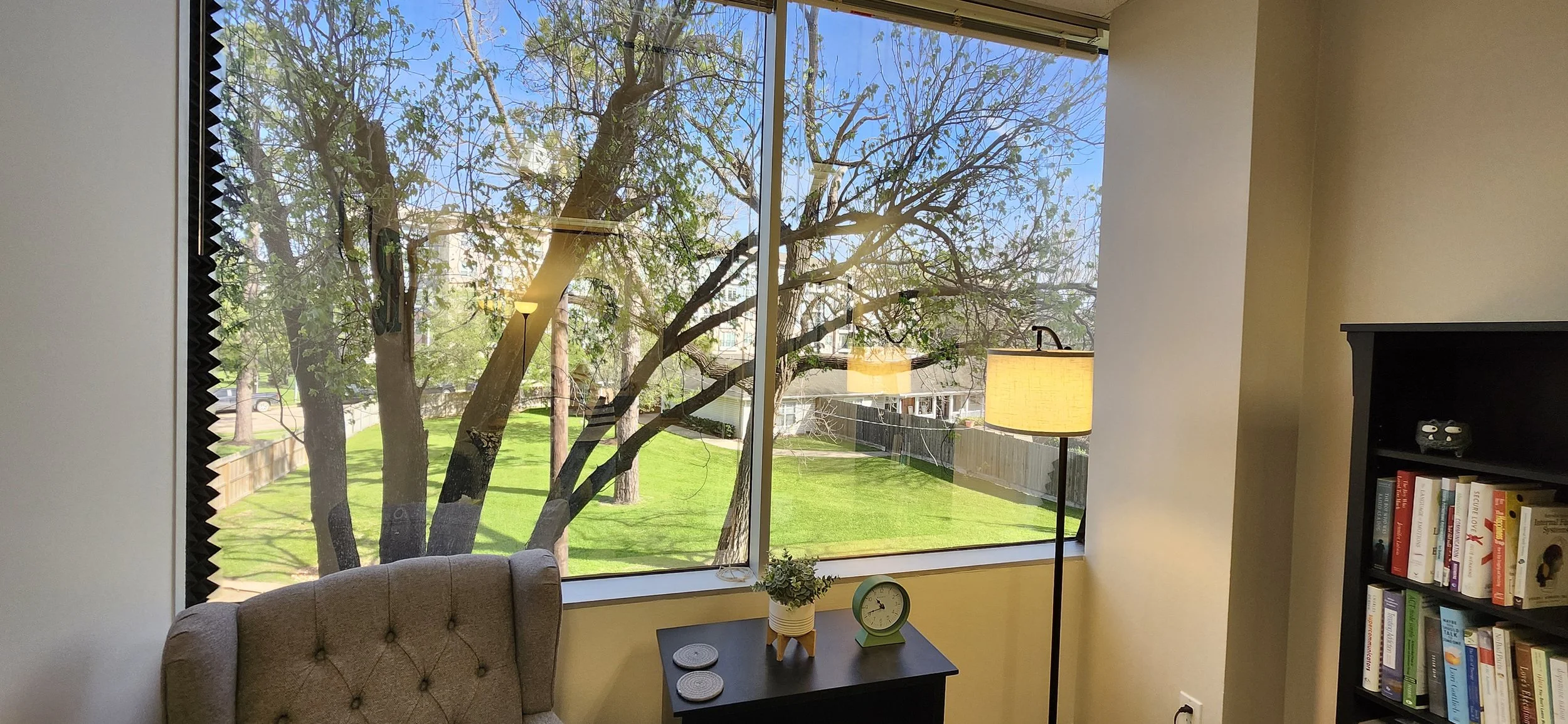 Living room with a large window showing a tree with green leaves and a grassy yard outside. Inside, there is a beige upholstered armchair, a black side table with a small potted plant, a green clock, and three coasters. A tall floor lamp with a beige